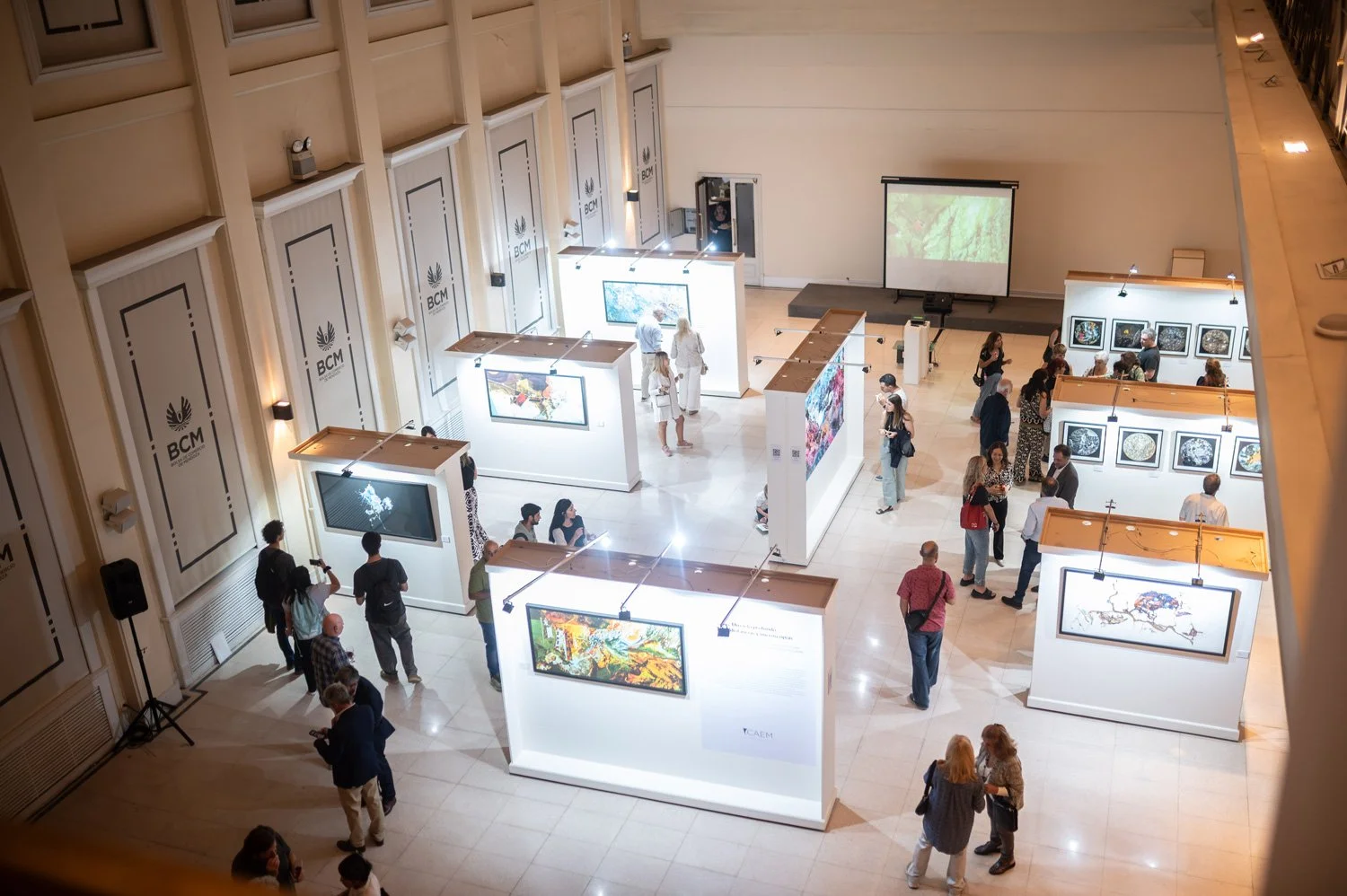 People viewing artwork in an art gallery with white walls and display panels, some engaging with digital screens and framed art pieces.