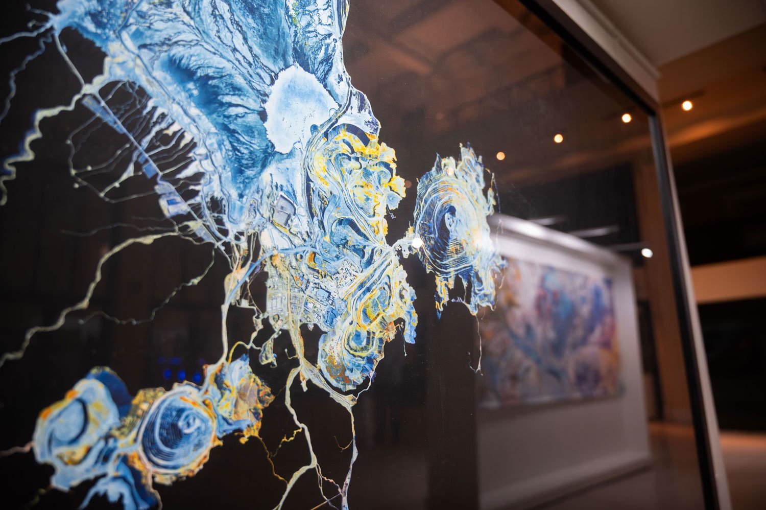 Abstract painting with swirling blue, yellow, and white patterns on a black background, displayed on a glass surface in an art gallery.