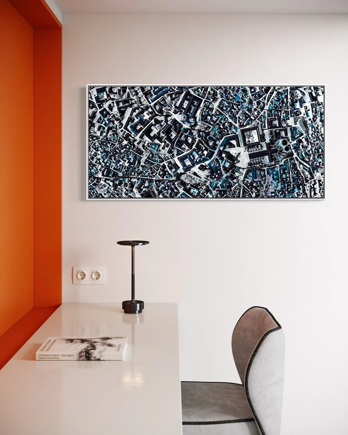 Modern office or living space with a white desk, a black lamp, a book, a gray chair, an electrical outlet, and a large abstract aerial map artwork on the wall.