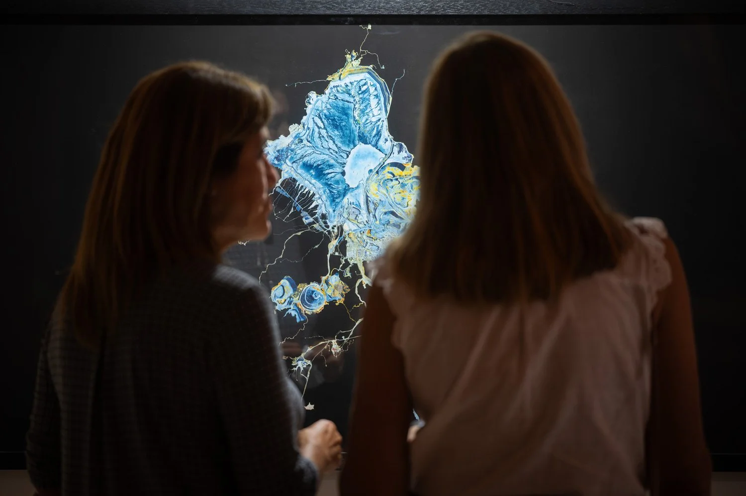 Two women observing a digital artwork or map with blue, white, and yellow details on a screen or display.