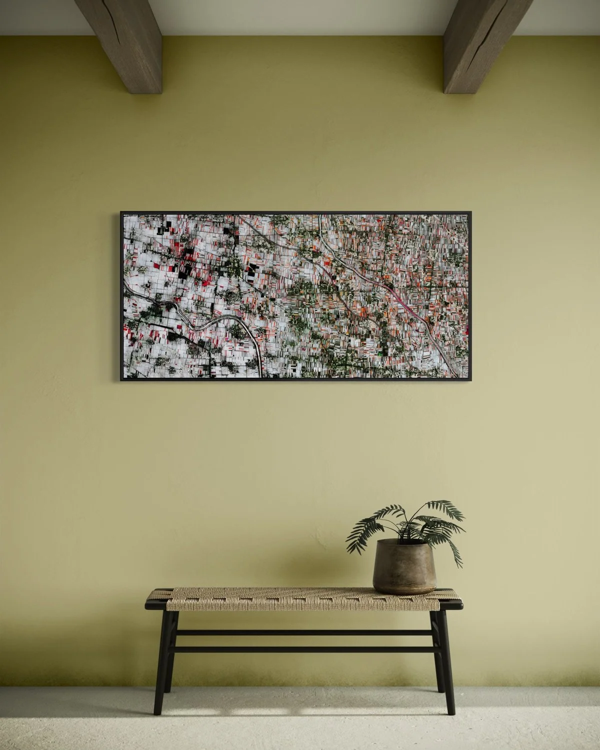 A minimalist room with a yellow wall, a framed aerial map, a bench with a woven seat, and a potted plant.