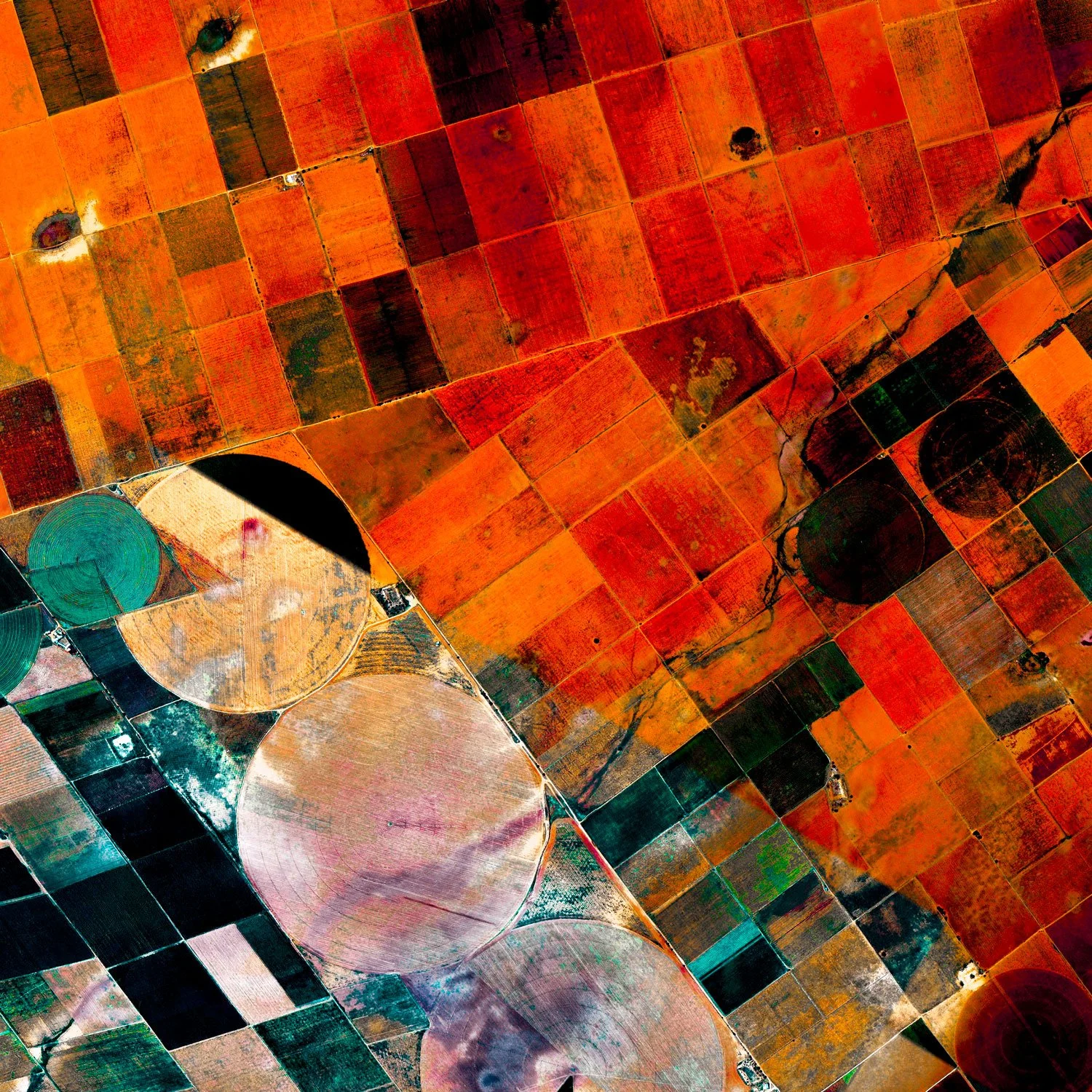 Aerial view of agricultural fields showing colorful patches of farmland, with cylindrical grain silos nearby.