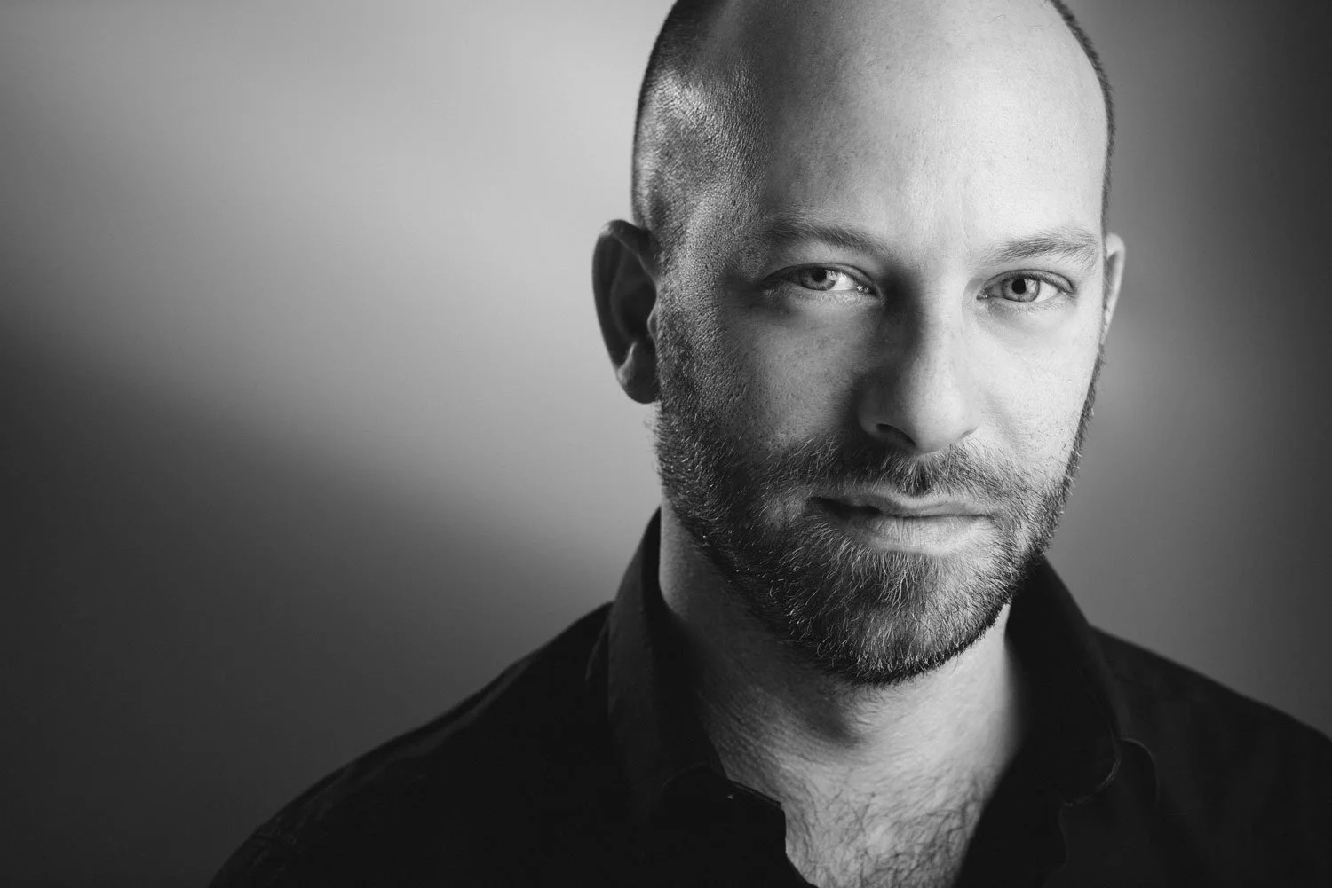 Black and white portrait of a man with light-colored eyes, a beard, and a shaved head, wearing a dark collared shirt against a plain background.