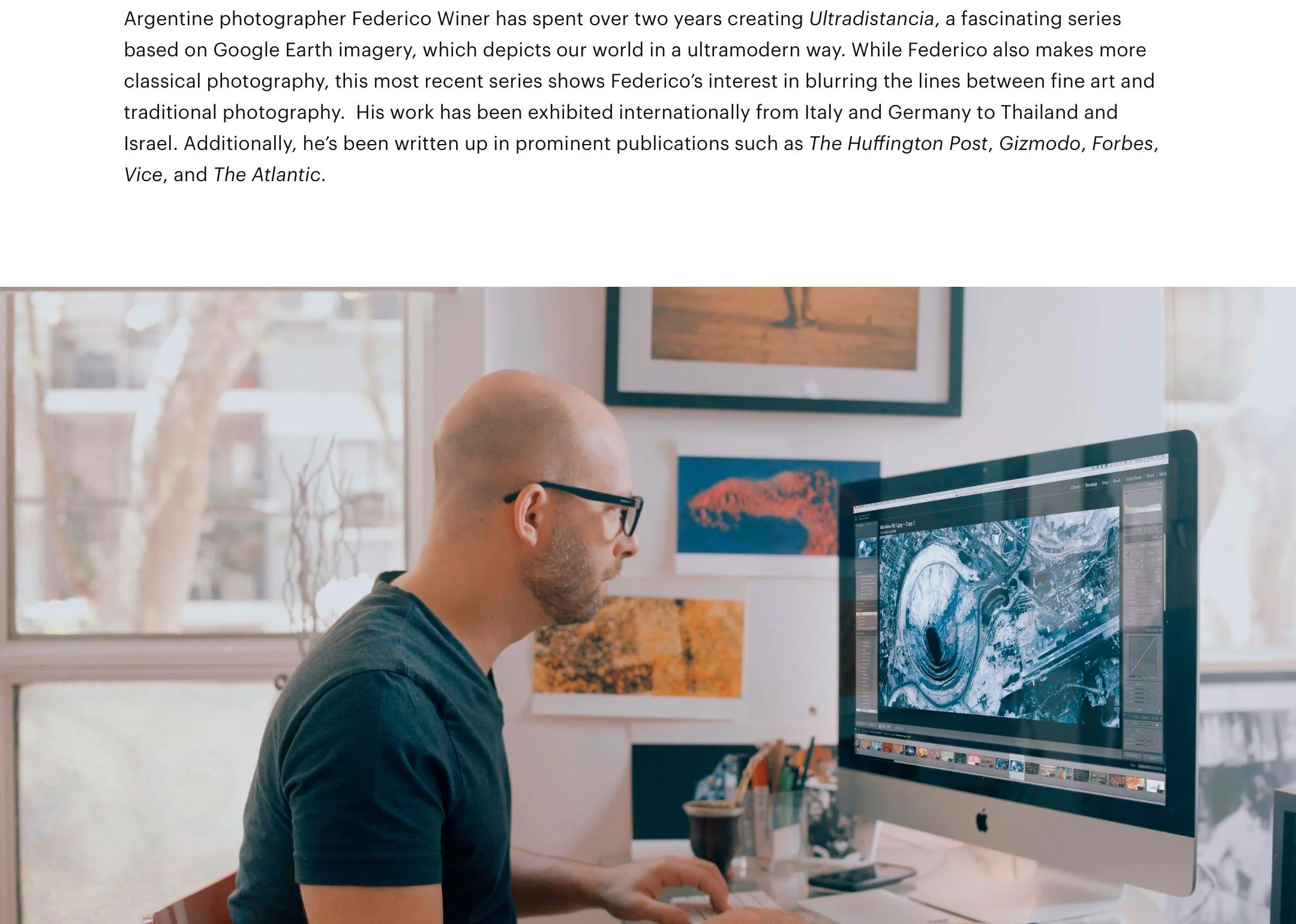 A man with glasses and a black shirt working on a computer with a large monitor displaying a digital artwork or map. The background shows framed pictures on the wall, a window with sunlight, and a few plants.
