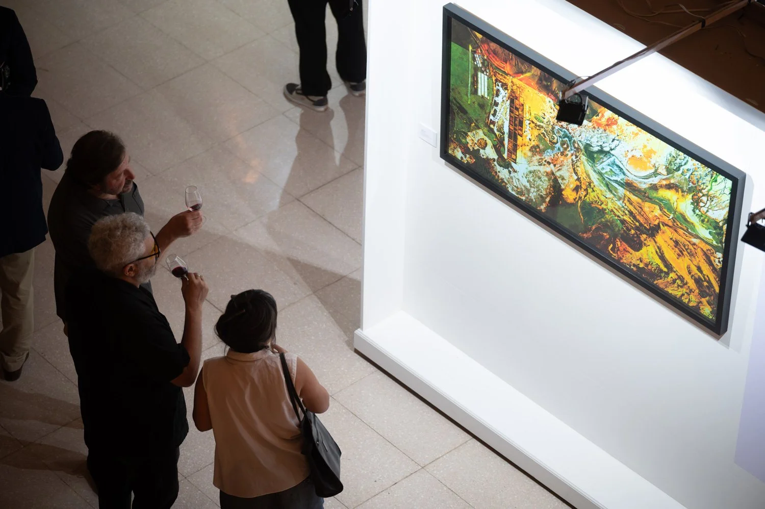 Three people in an art gallery holding glasses of red wine, looking at a colorful landscape painting on a TV screen mounted on a white display wall.