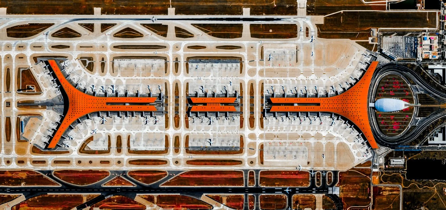 Aerial view of Beijing Daxing International Airport, showing its futuristic, star-shaped design with orange inlaid sections and surrounding runways and taxiways.
