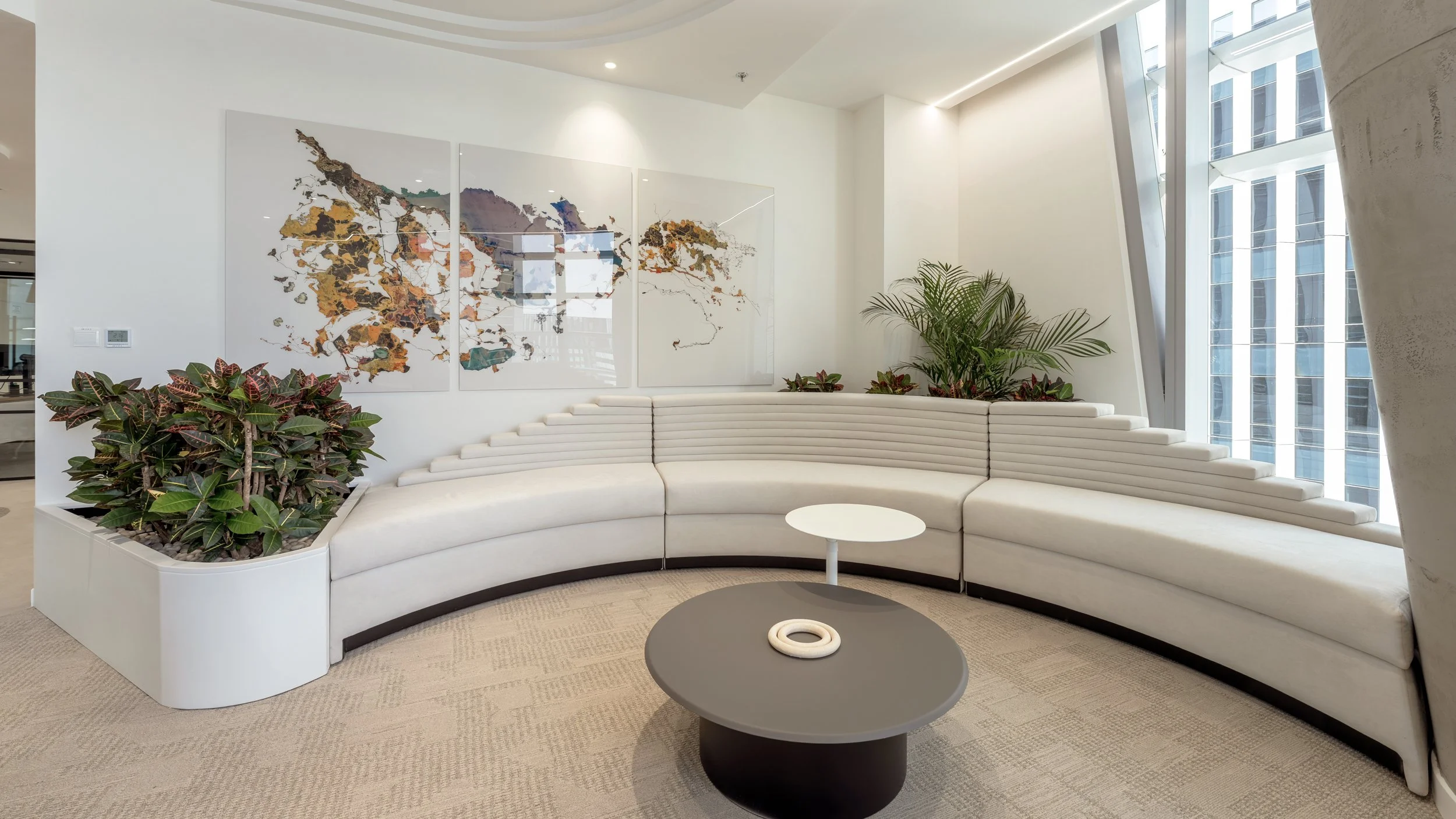Modern lobby with a white curved seating area, indoor plants in a white planter, abstract world map artwork on the wall, large windows, and a small coffee table.