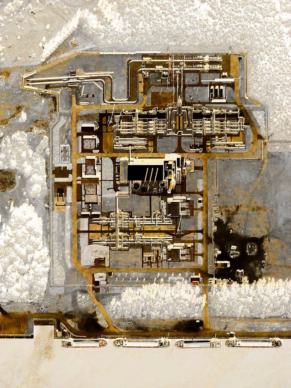 An aerial view of a large industrial complex with multiple buildings, pipelines, and roads, surrounded by snow and barren landscape.