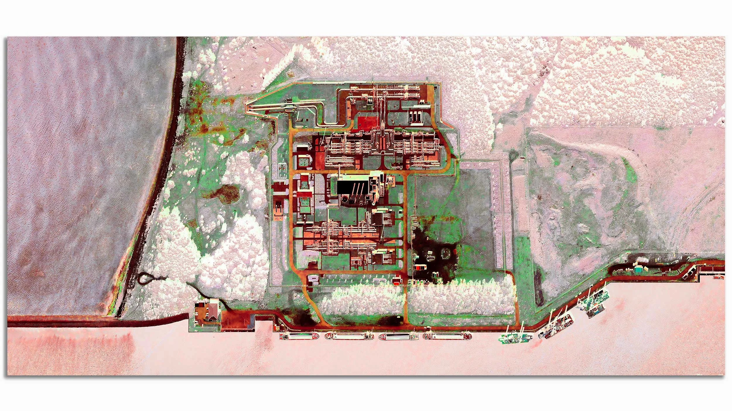 Aerial view of a large industrial facility or power plant with multiple buildings, piping, and infrastructure, surrounded by a fence and adjacent to a body of water.