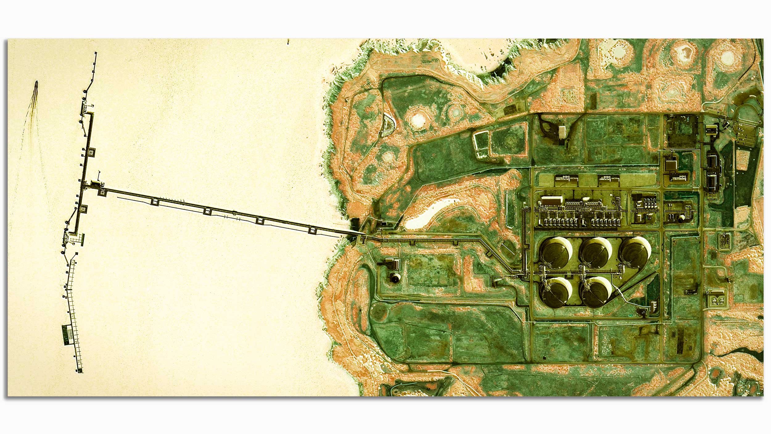 Aerial view of a large industrial site with tanks, buildings, and machinery, adjacent to a landscape with roads and green spaces.