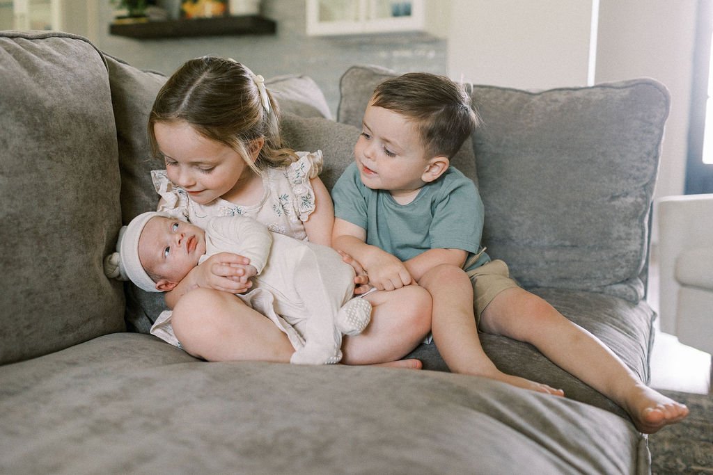 A boy and girl toddler hold their younger newborn brother on the couch.