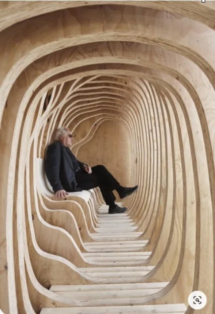 Person sitting inside a tunnel-like wooden structure with curved, layered walls.