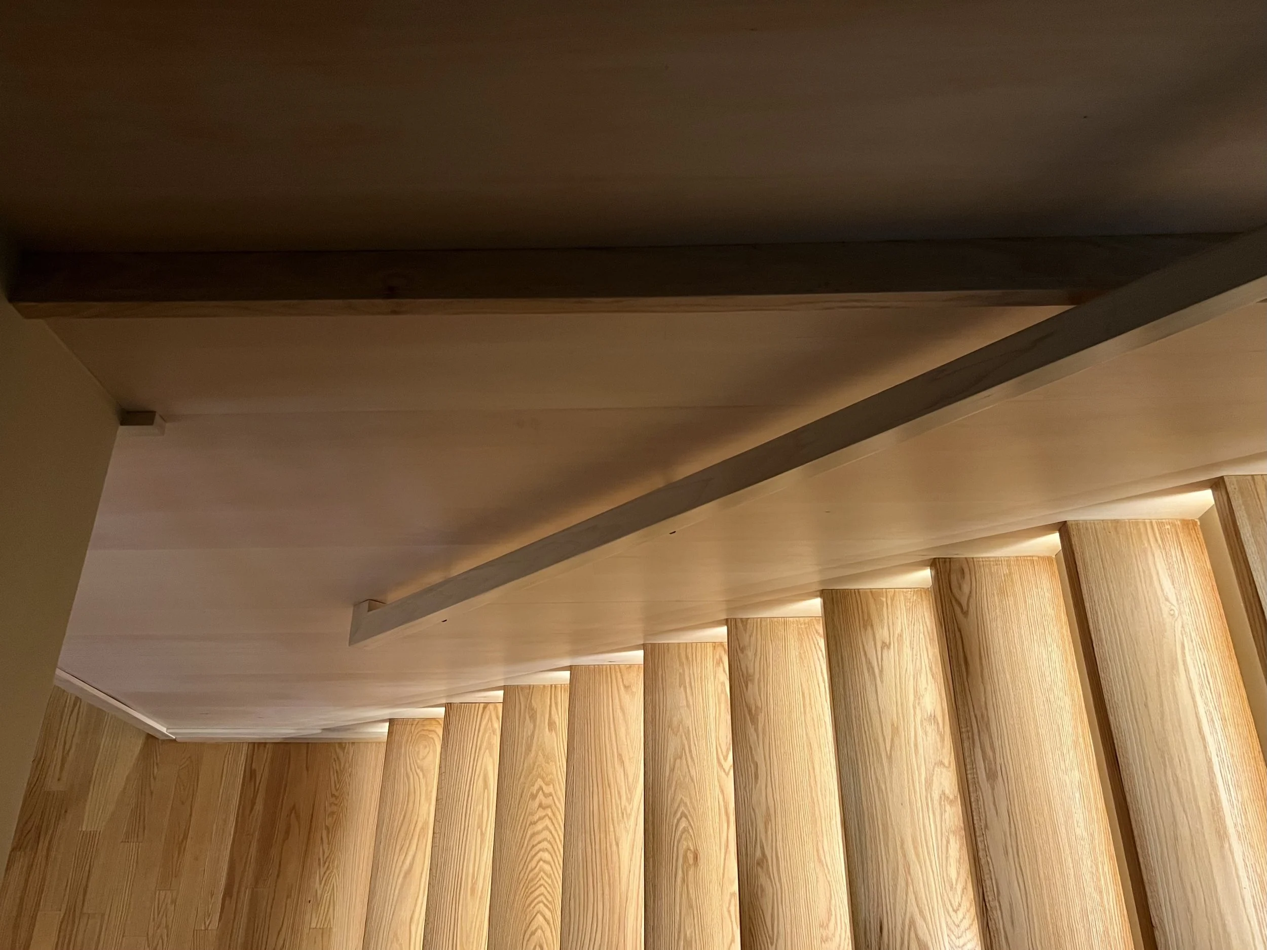 Wooden staircase with light-colored wood and a handrail on the right side, illuminated by warm lighting under every step.