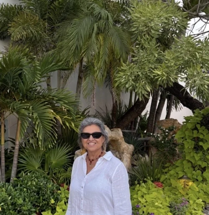 Smiling woman wearing sunglasses and white shirt standing in lush tropical garden with various green plants and trees.