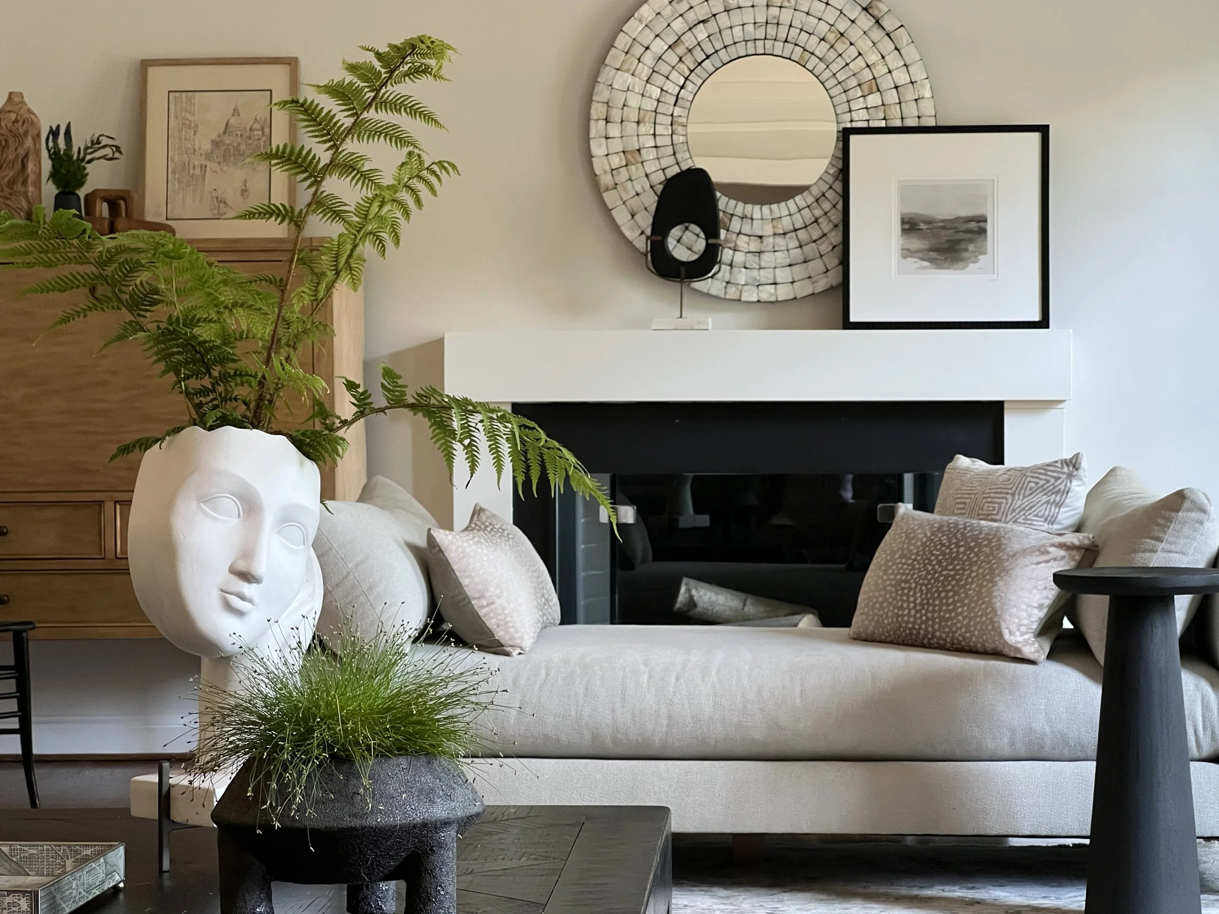 Living room with beige lounge daybed for the front of fireplace, decorative pillows, large green fern in a white face-shaped planter, black coffee table; wall art, round mirror, framed black-and-white landscape photo on a modern fireplace-mantel.