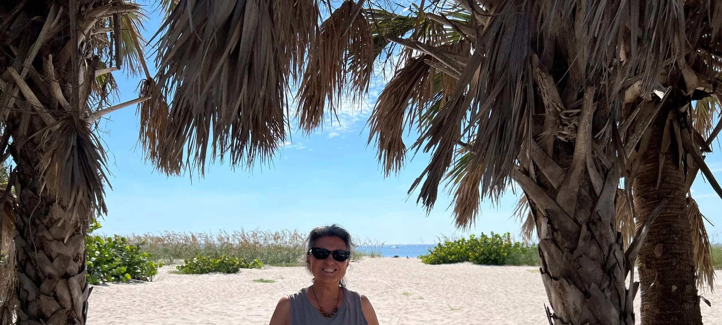 Smiling woman in sunglasses sitting on a beach under a large palm tree with the ocean and blue sky in the background.