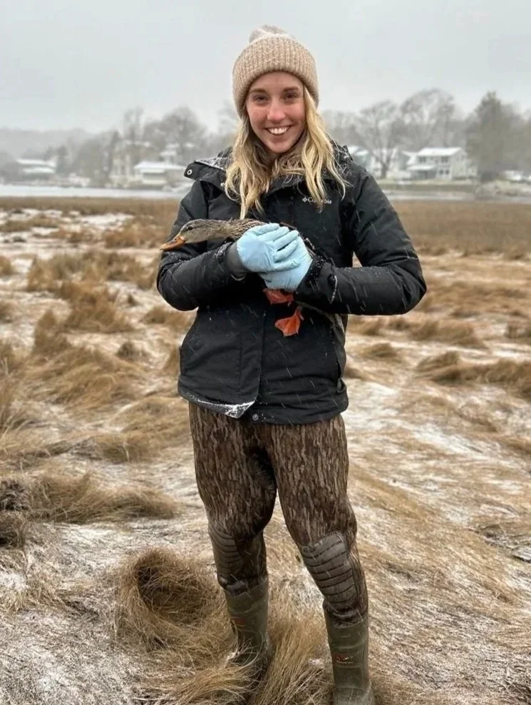 A woman outdoors in a snowy field holding a small fish with both hands, smiling at the camera. She's wearing a beige knit hat, a black winter jacket, camo-patterned pants, and green rubber boots.