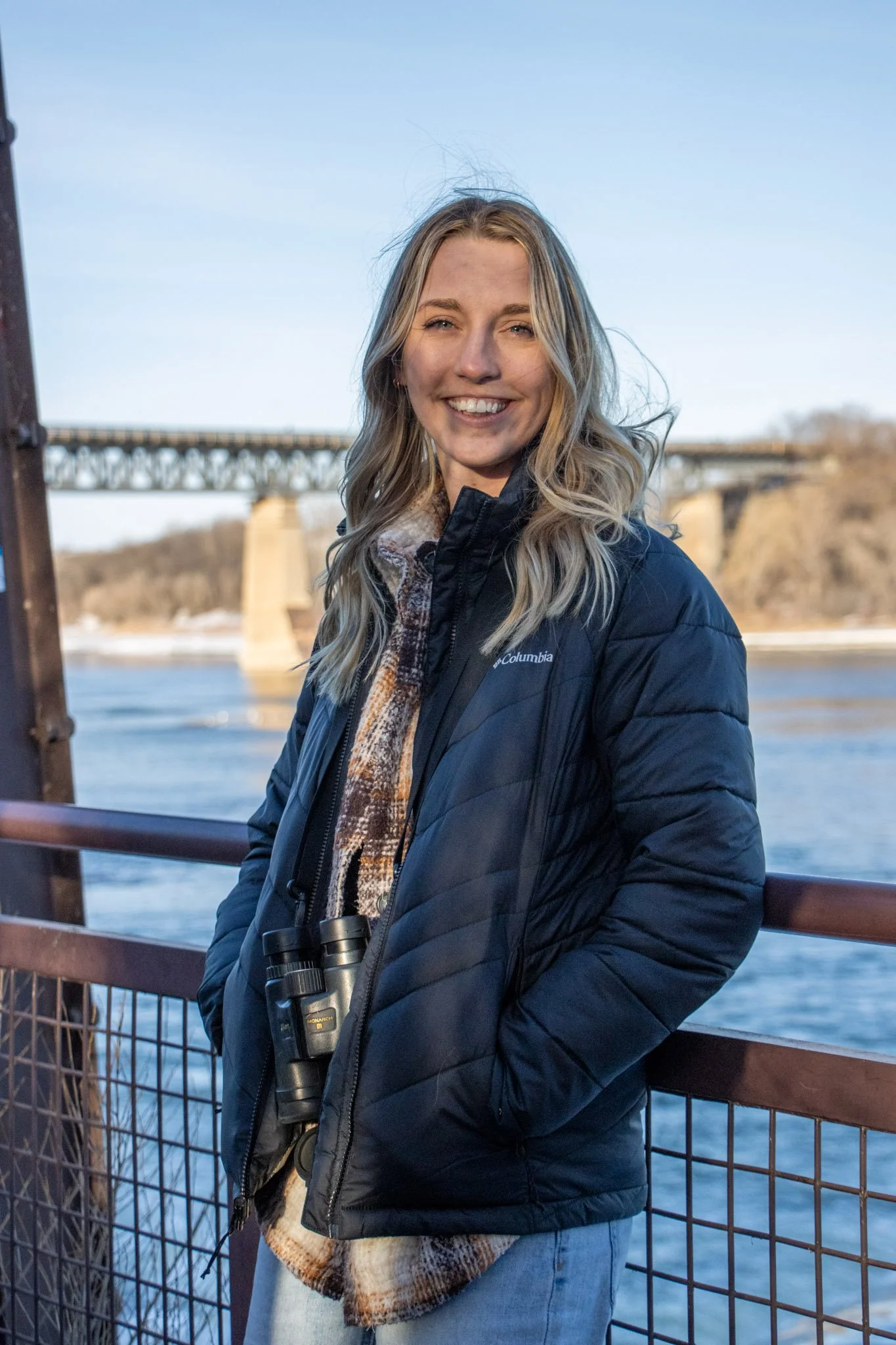 A young woman with long blonde hair smiling while standing on a bridge overlooking a river, wearing a navy Columbia jacket, a scarf, and binoculars around her neck.