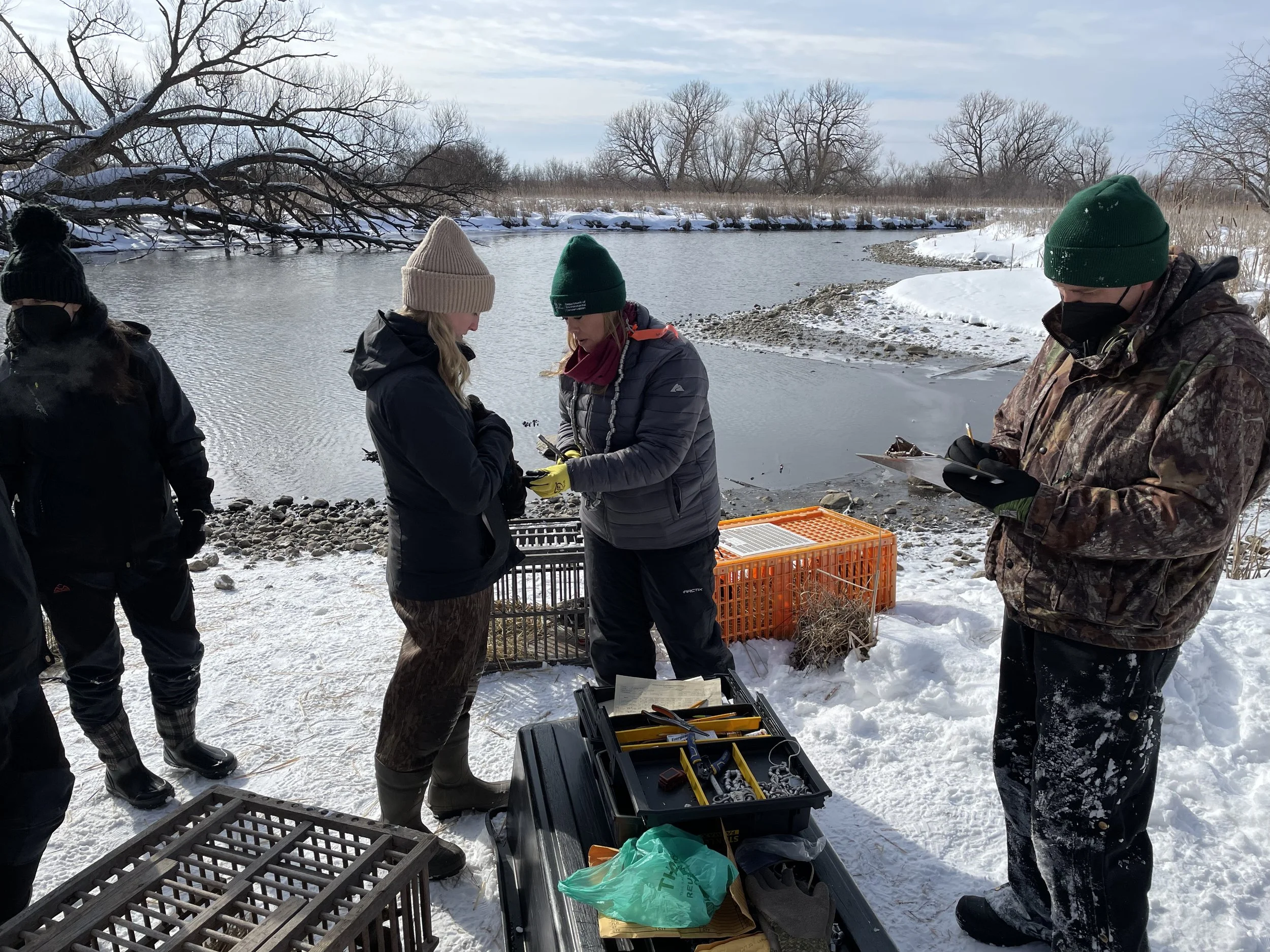 A group of people in winter gear conducting fieldwork at a snowy riverbank, with equipment and containers nearby.