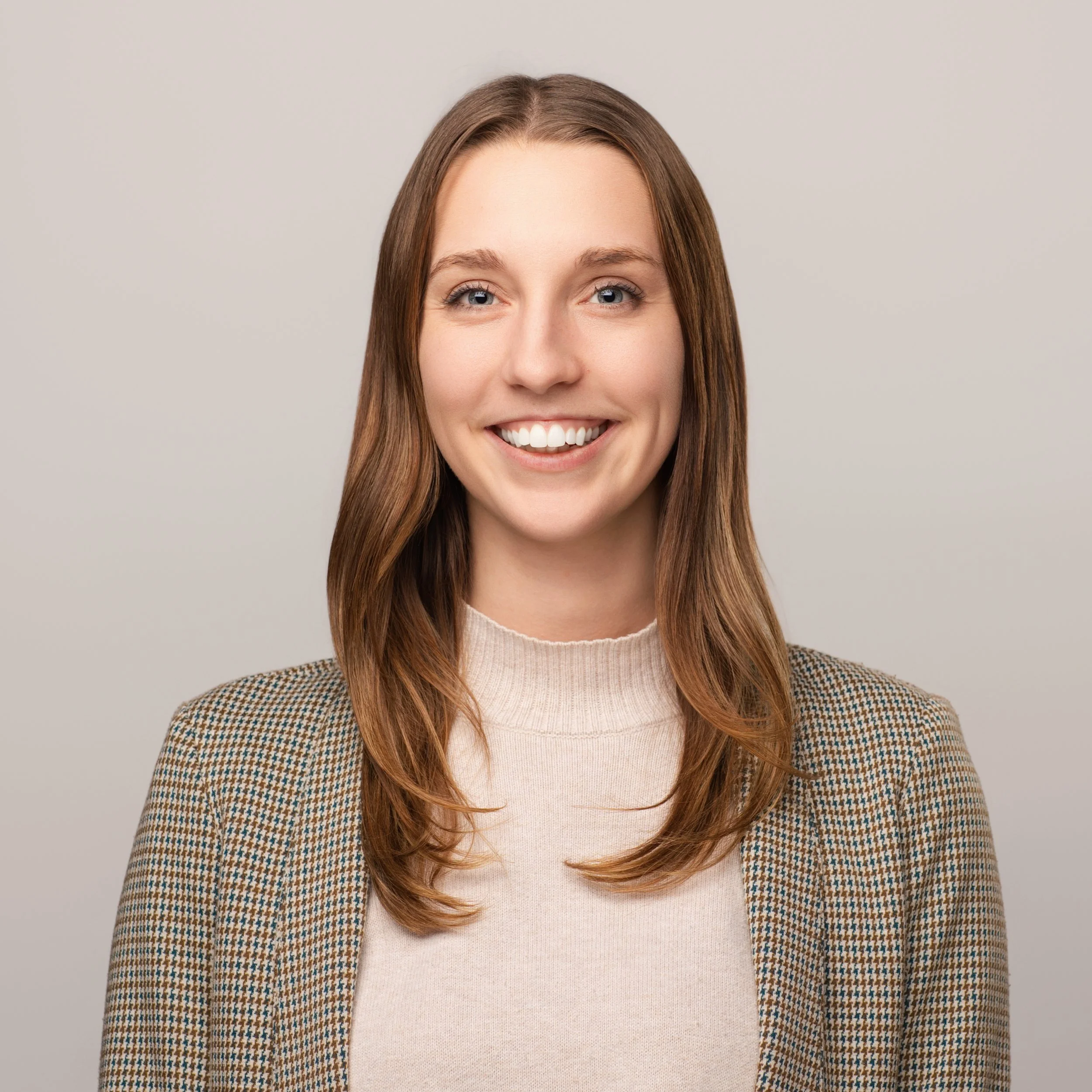 A young woman with long brown hair smiling, wearing a beige turtleneck and a checked blazer, against a plain light background.