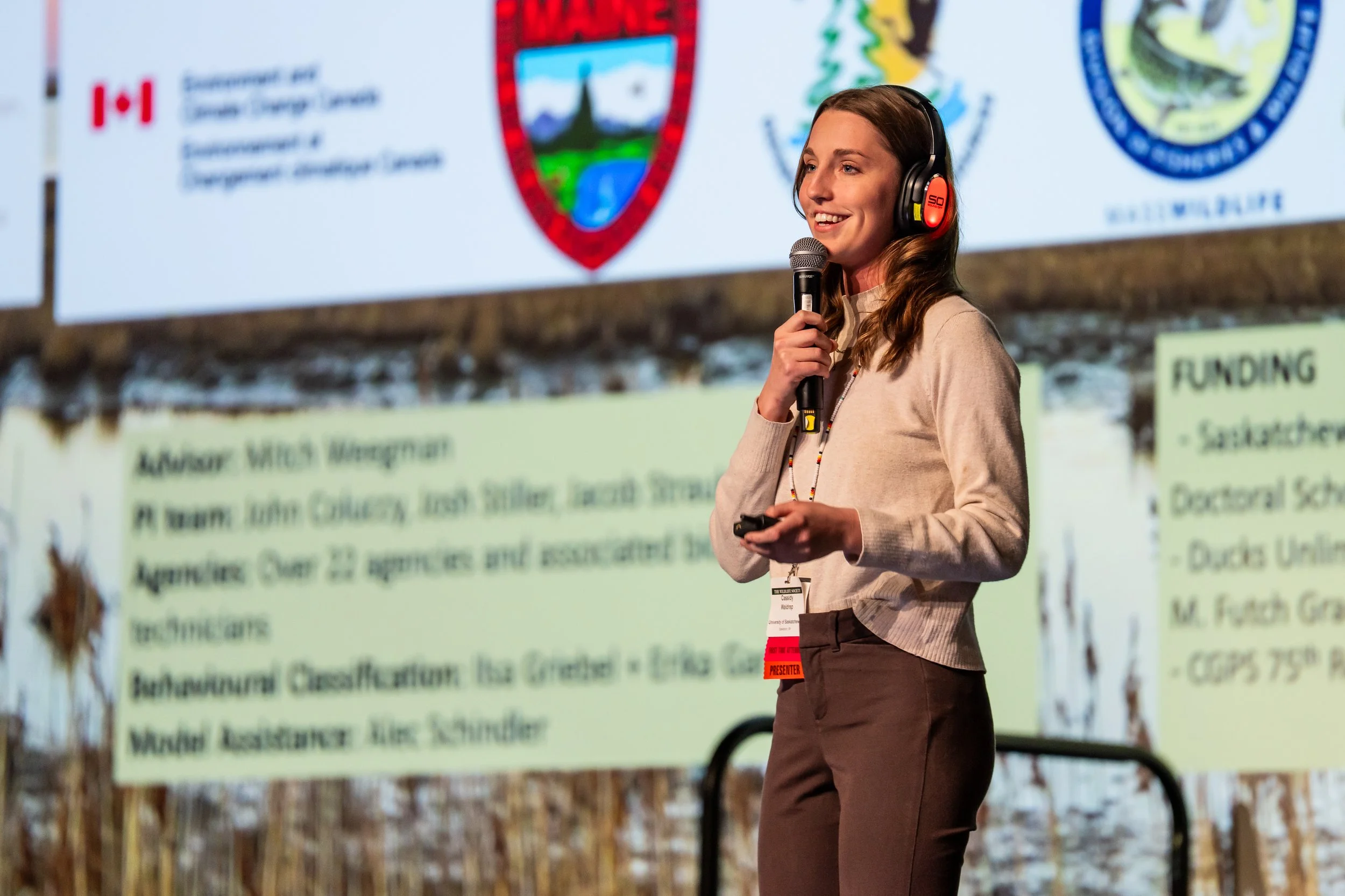 Woman speaking at a conference or seminar with a microphone, wearing headphones, in front of a screen displaying logos of Canadian government agencies and a presentation slide with text.