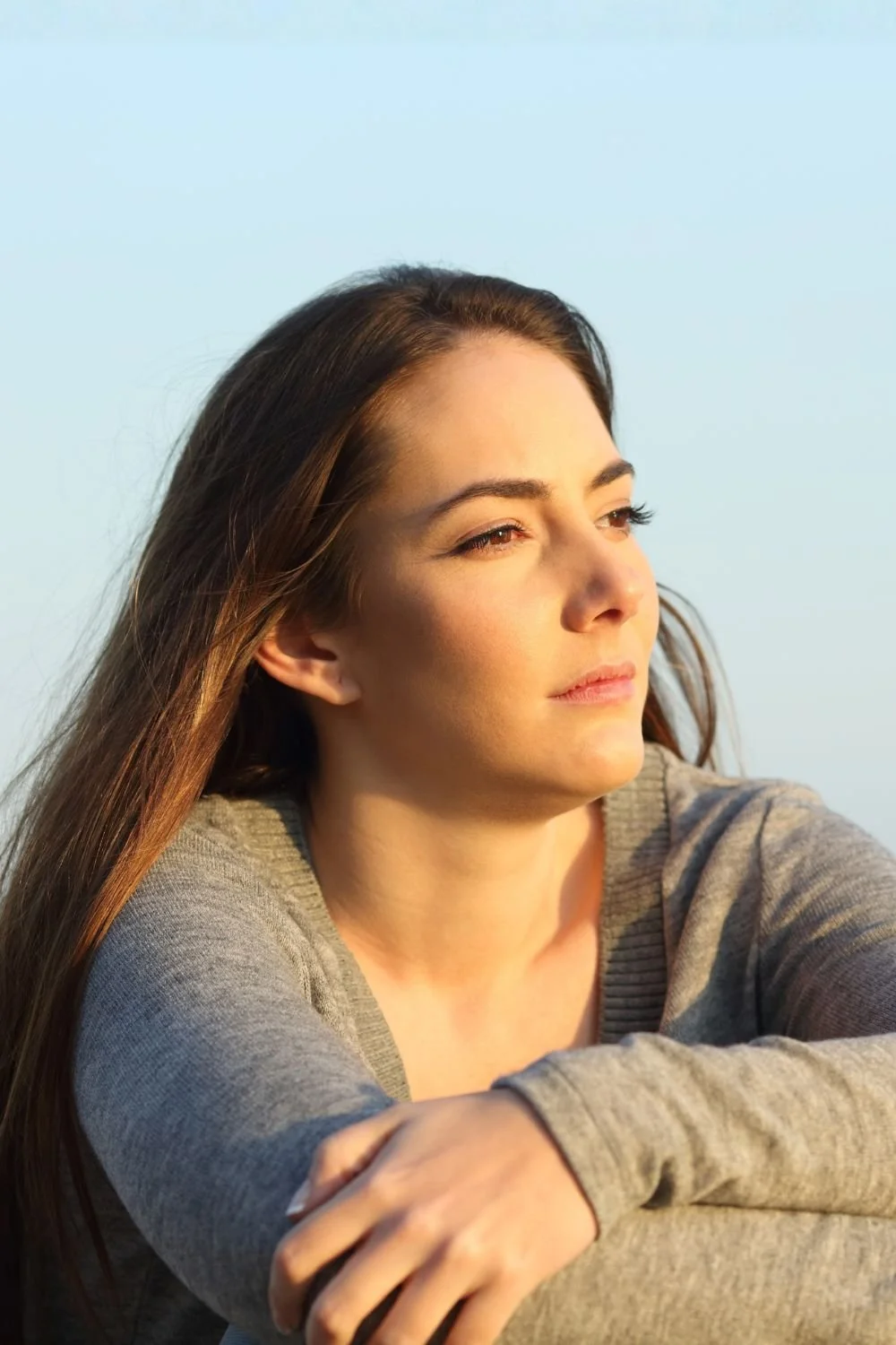A woman with long brown hair and fair skin gazing into the distance during daylight with a clear sky in the background.