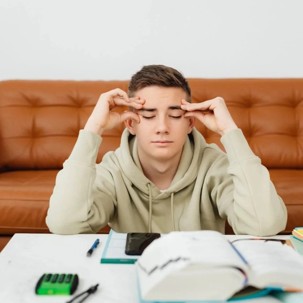 A young man with short brown hair sitting at a table with school supplies, holding his head and appearing stressed or overwhelmed.