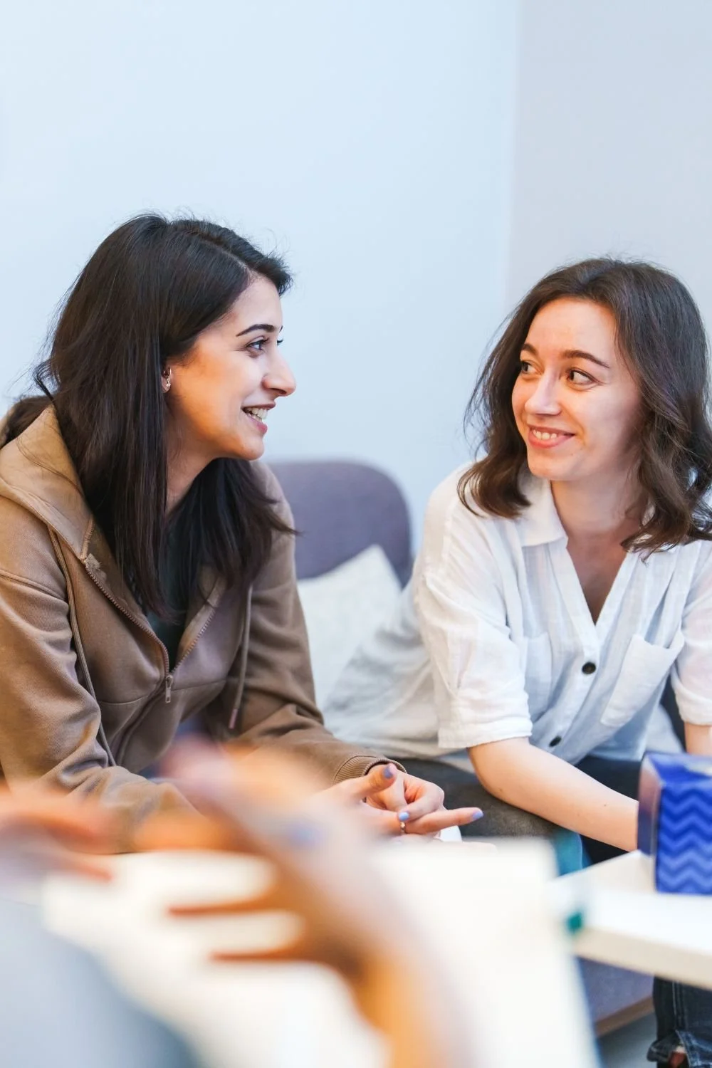 Two women sitting and talking, smiling at each other, in a casual indoor setting.