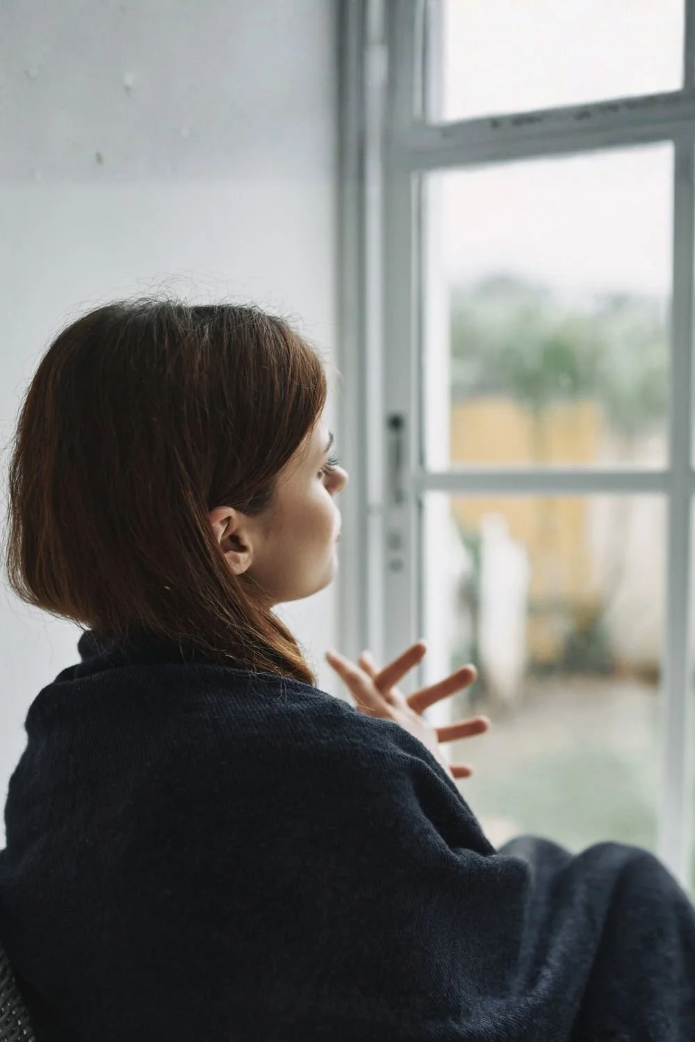 A woman with short brown hair gazes out a window, touching the glass with one hand, in a softly lit room.