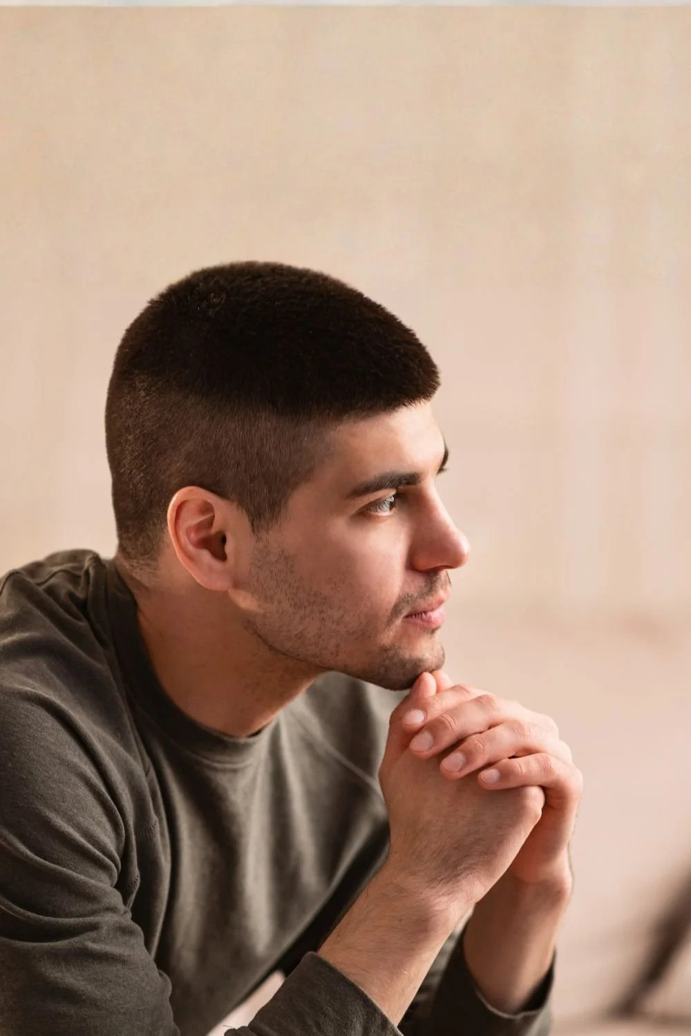 Side profile of a young man with short dark hair and light stubble, resting his chin on his clasped hands, looking thoughtful.
