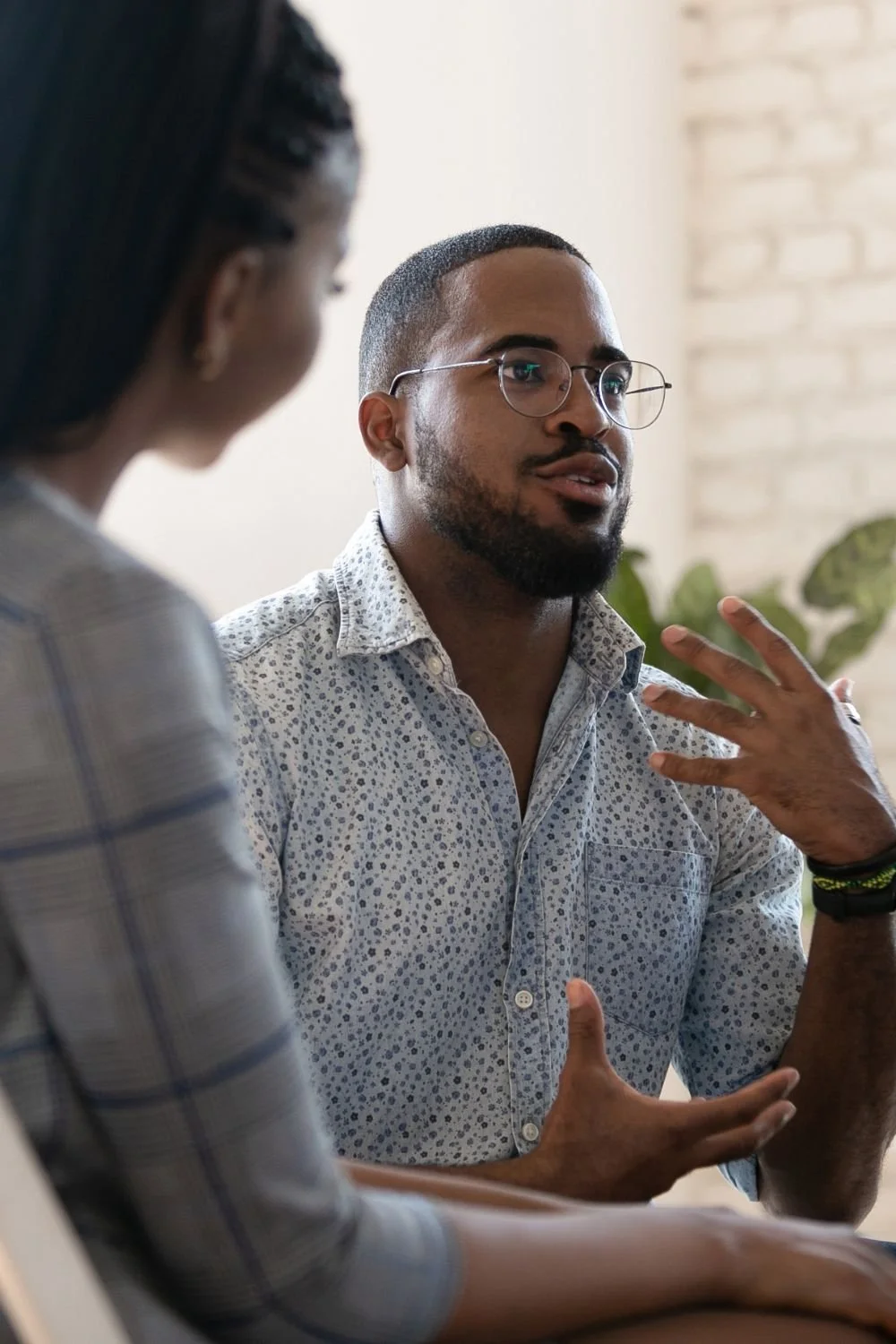 A man with glasses and a patterned shirt having a conversation with a woman in a plaid shirt, in an indoor setting with a plant and a brick wall in the background.