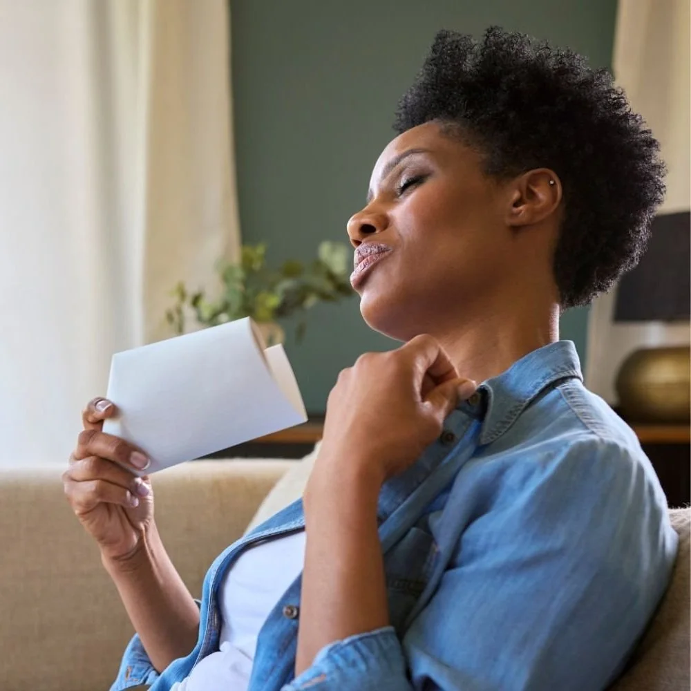 woman on couch in blue shirt waving paper to cool down