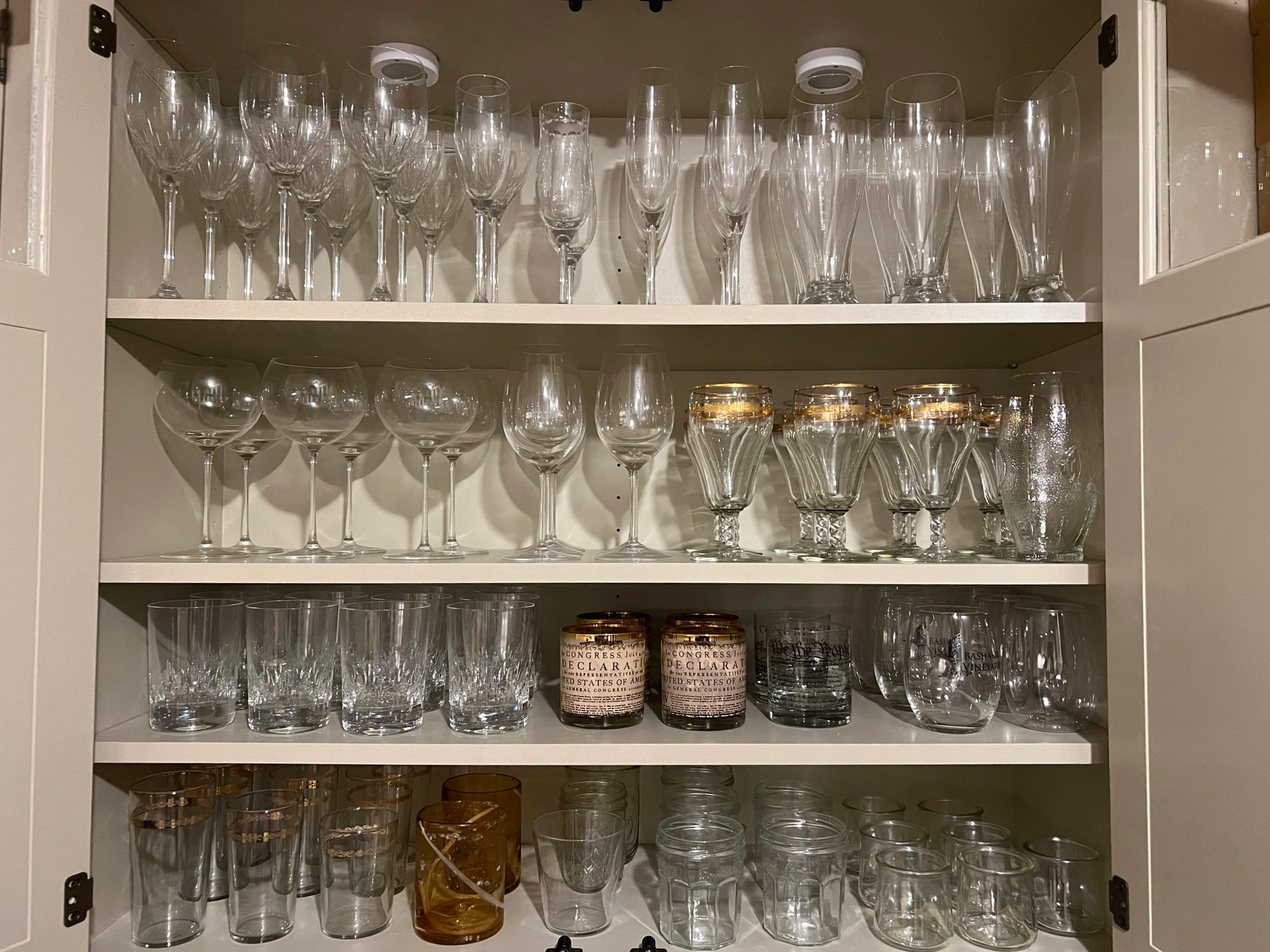 A cabinet filled with various types of glassware, including wine glasses, cocktail glasses, and tumblers, organized on four shelves.