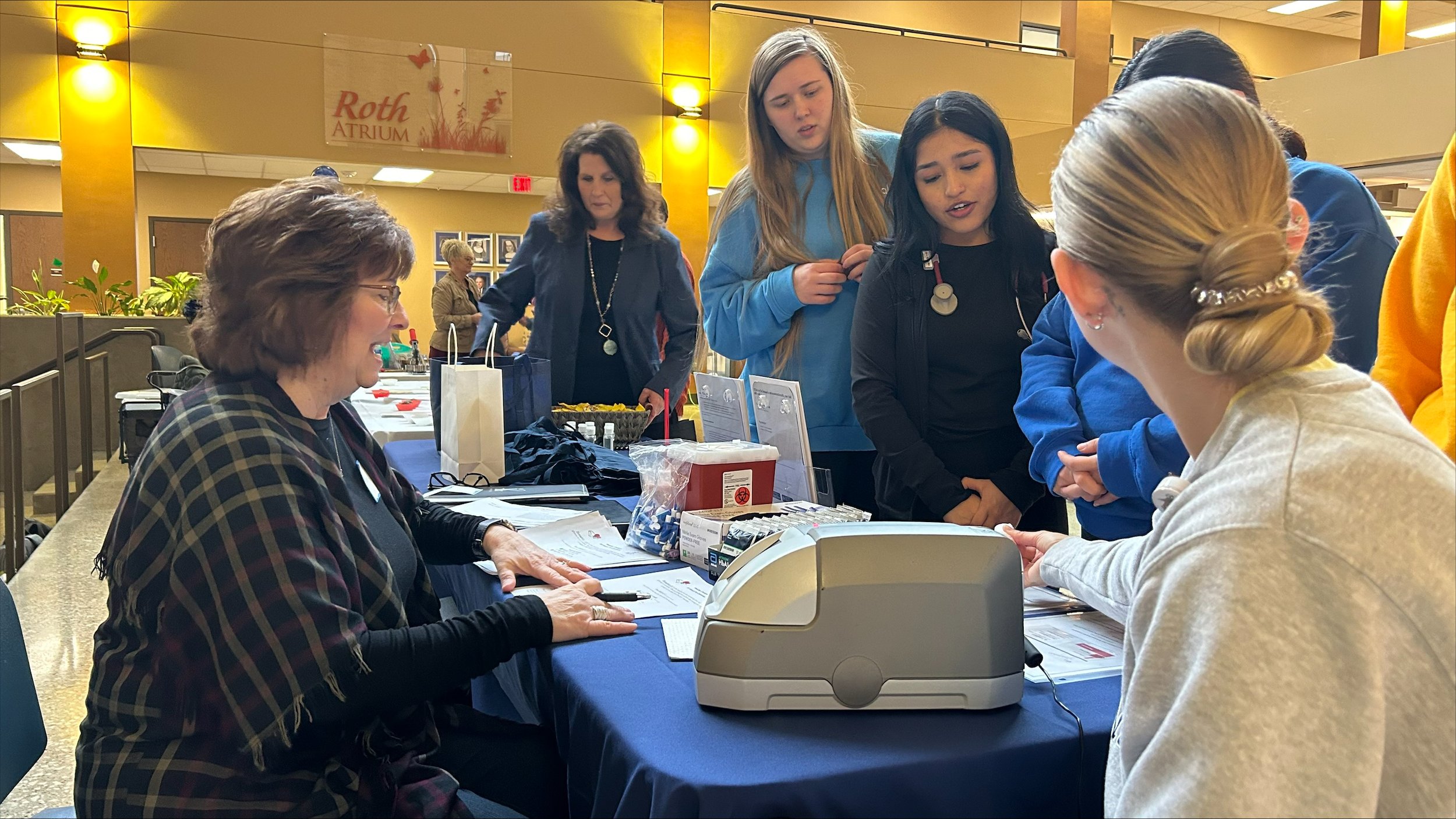  BCU students crowd around the IBC Innovative Business Consultants booth, while Veronica Pitzl got the opportunity to test her HbA1C (Hemoglobin A1c) via a fingerstick, a frontline test for Diabetes.&nbsp;&nbsp; 