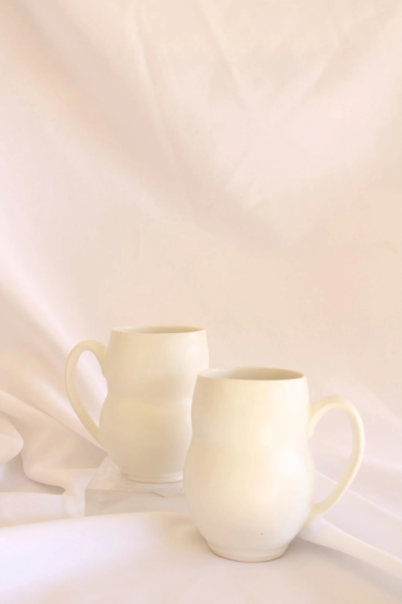 Two cream-colored ceramic mugs with handles, placed on a soft, light-colored fabric background.