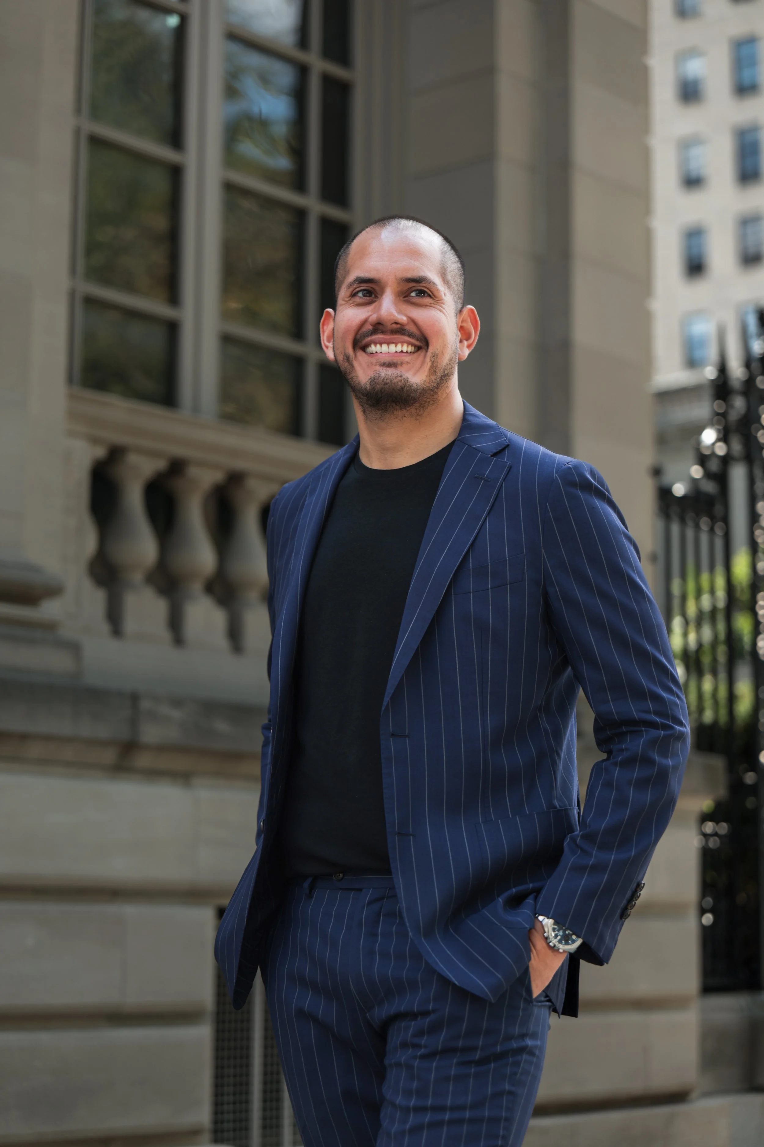 A man with a shaved head and a beard, smiling and dressed in a blue pinstripe suit with a black shirt, standing outdoors in front of a building with large windows and a black fence.