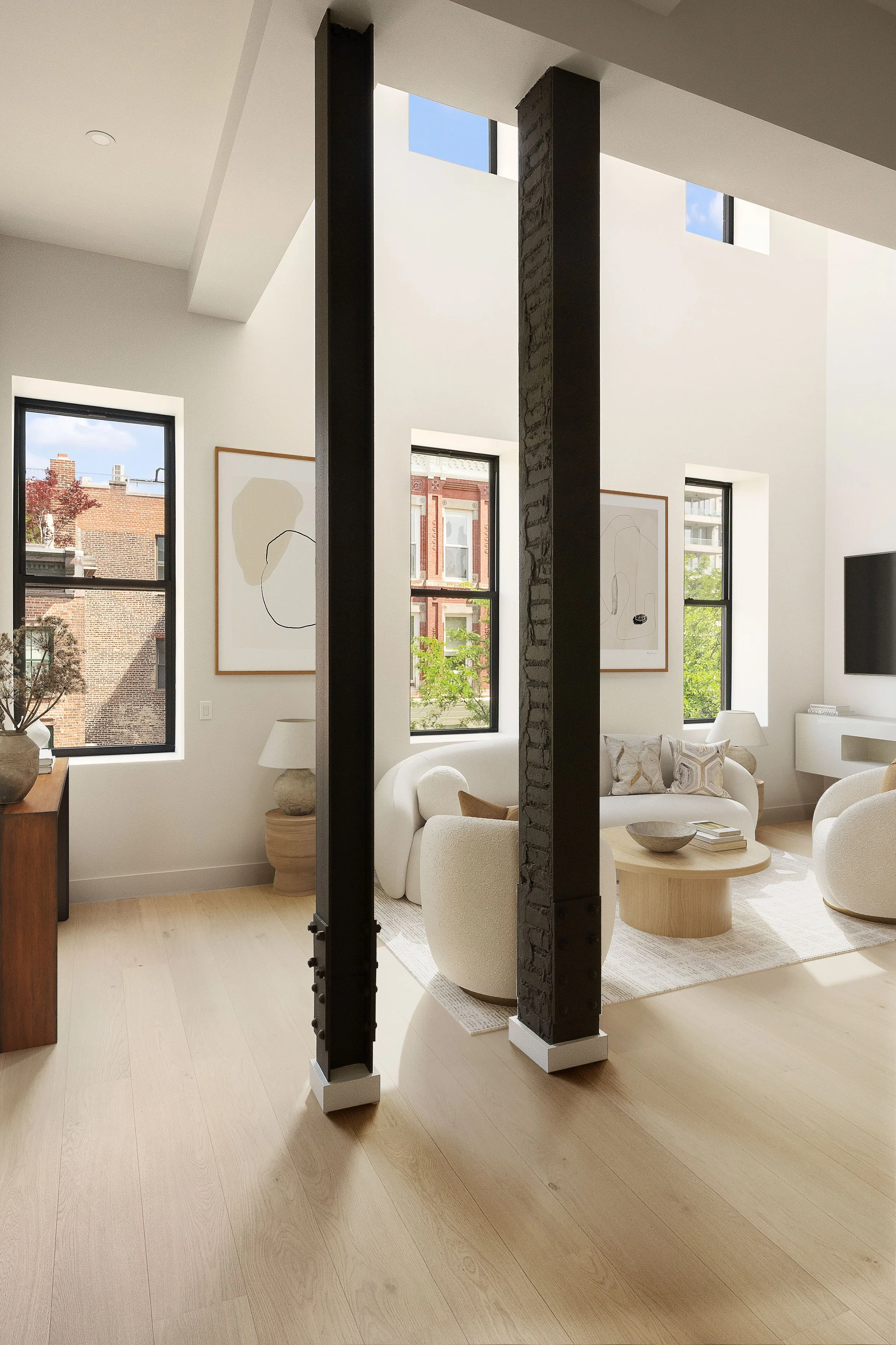 Modern living room with large windows, white walls, and contemporary furniture including a white sofa, armchair, and round coffee table. Artwork hangs on the walls, and natural light enhances the space's airy feel.