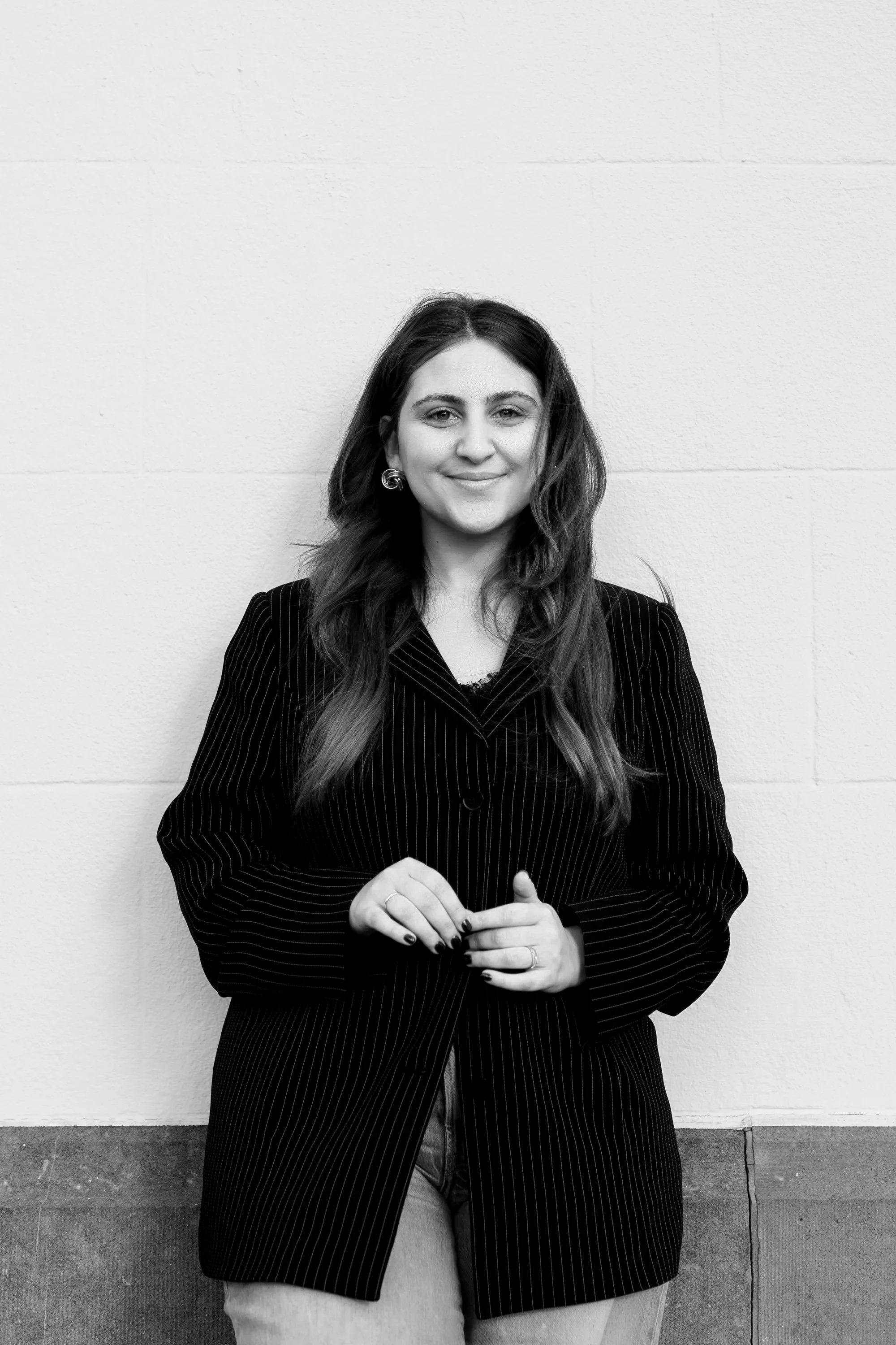 Zwart-wit foto van een lachende vrouw met lang haar, gekleed in een pinstripe blazer, staand tegen een witte muur.
