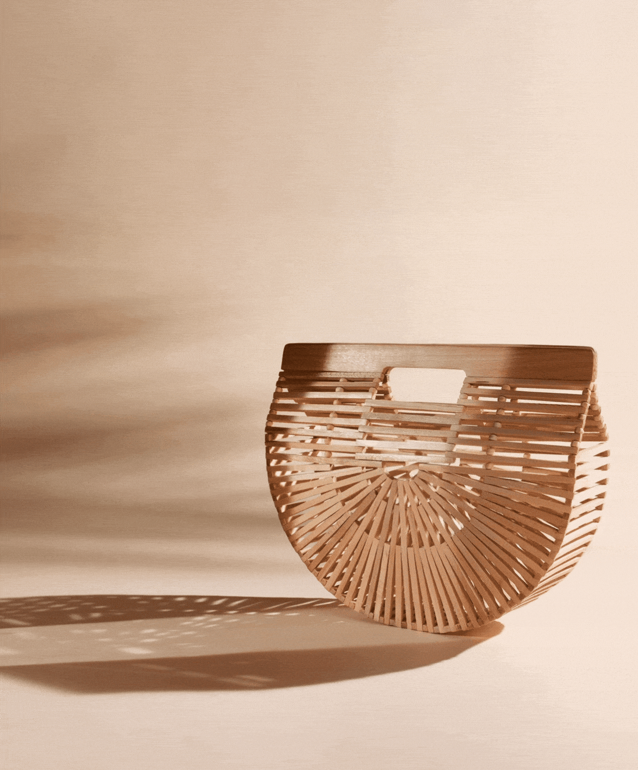 A modern, semi-circular wooden handbag with a slatted design and cut-out handle, casting a shadow on a light beige surface.