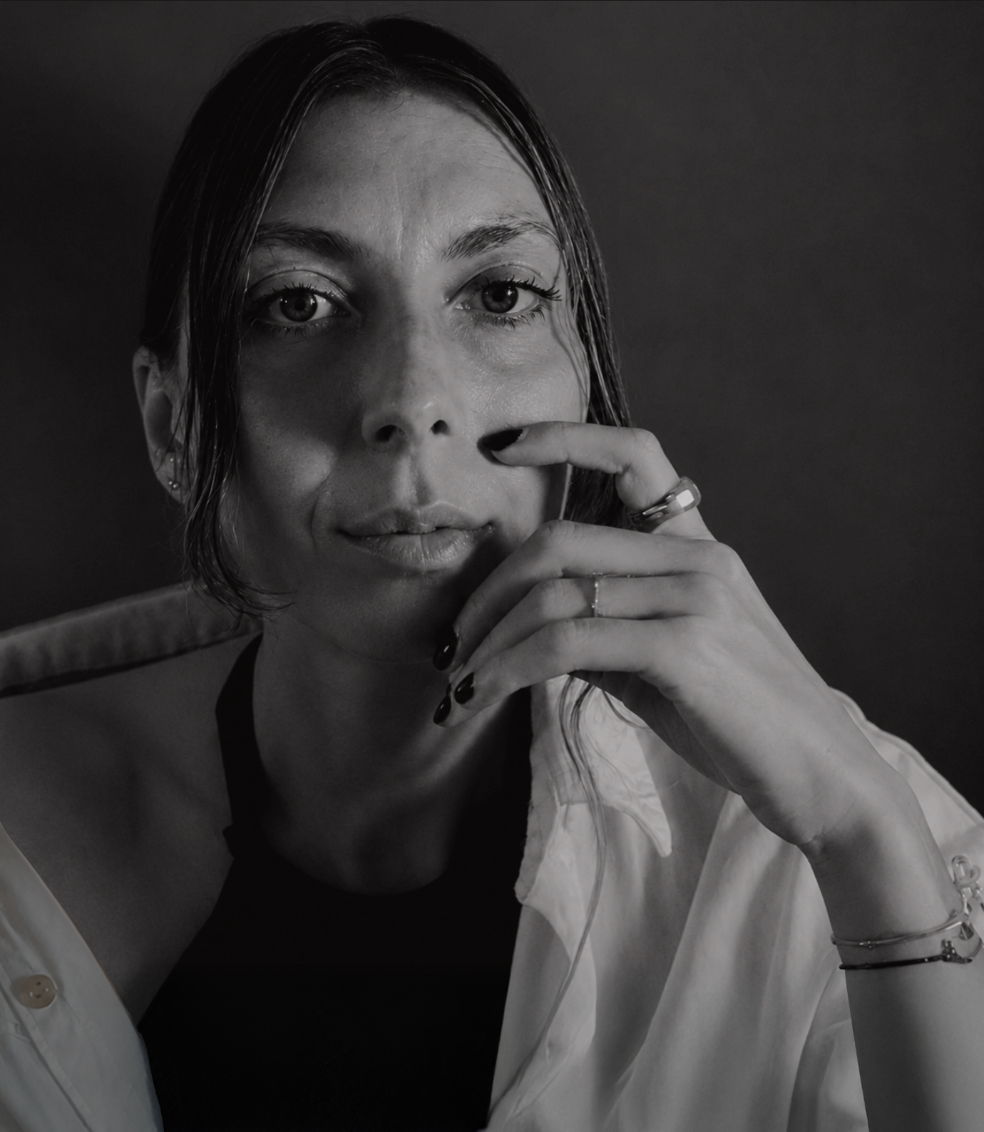 Black and white portrait of a woman with wet hair, touching her face with her right hand, wearing rings and earrings, against a dark background.