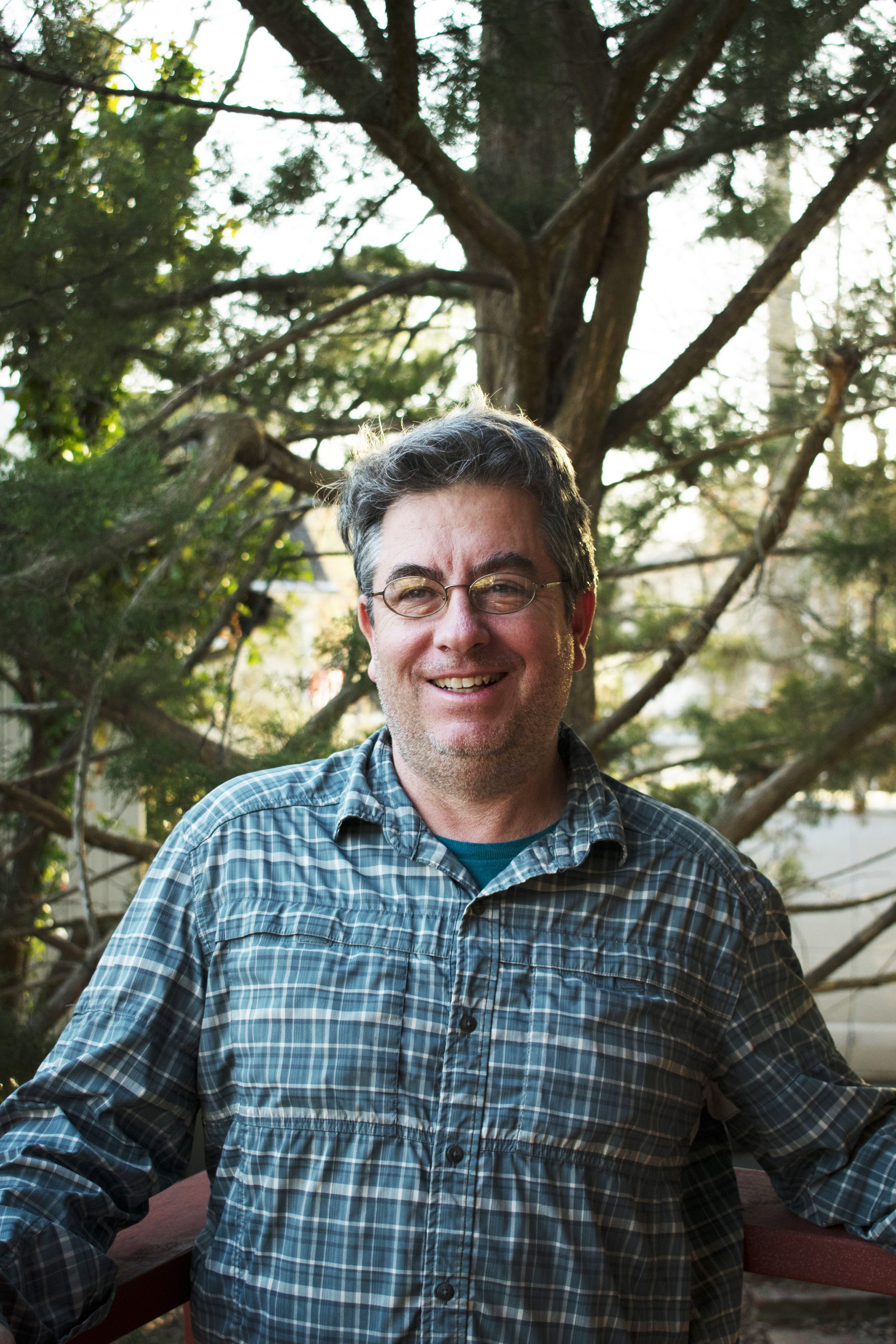 Paul Sturm, smiling in front of trees, wearing a blue checkered shirt and glasses