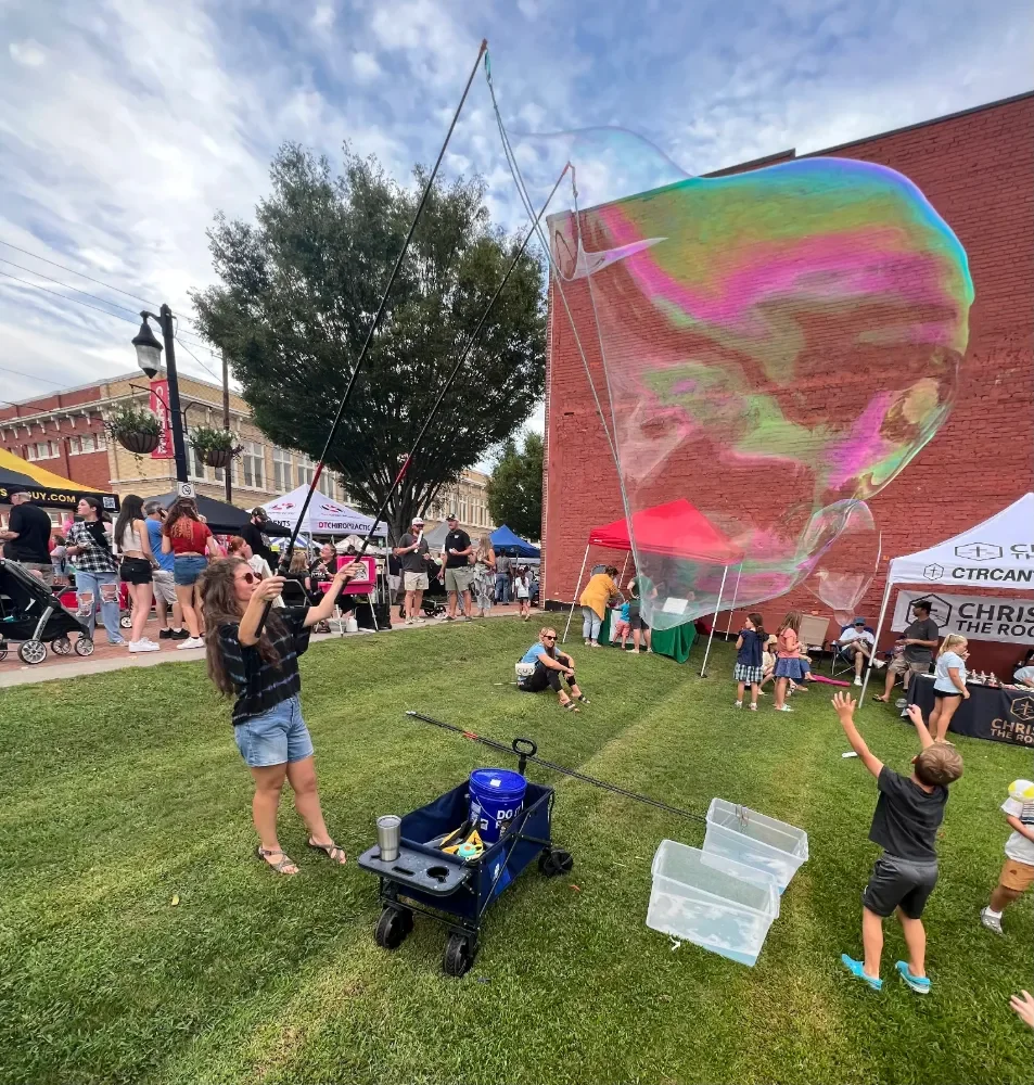 A Bubble Vision Atlanta team member creating a large soap bubble at a festival or fair with many people, tents, and booths in the background.