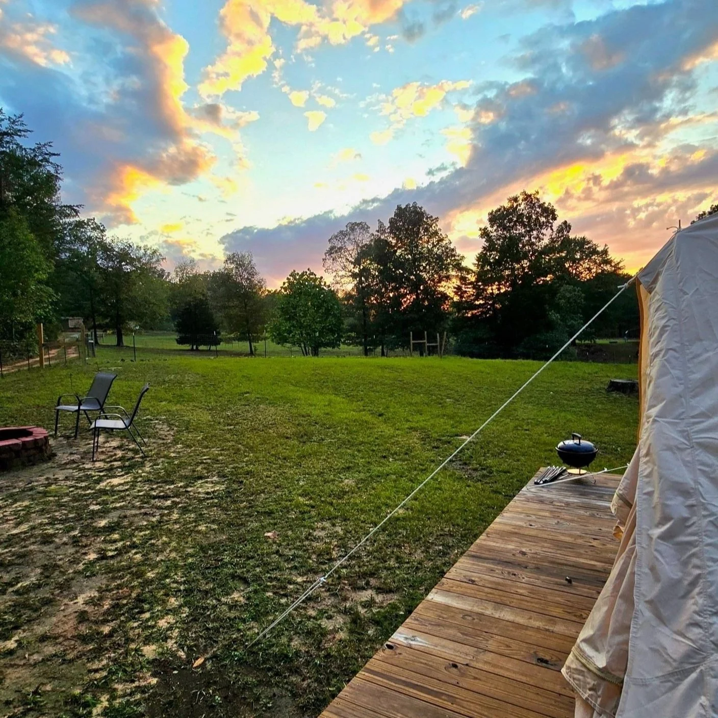 View of a backyard during sunset with a grassy lawn, trees in the background, two chairs on the left, a fire pit, a partially visible tent on the right, and a small grill on the deck.