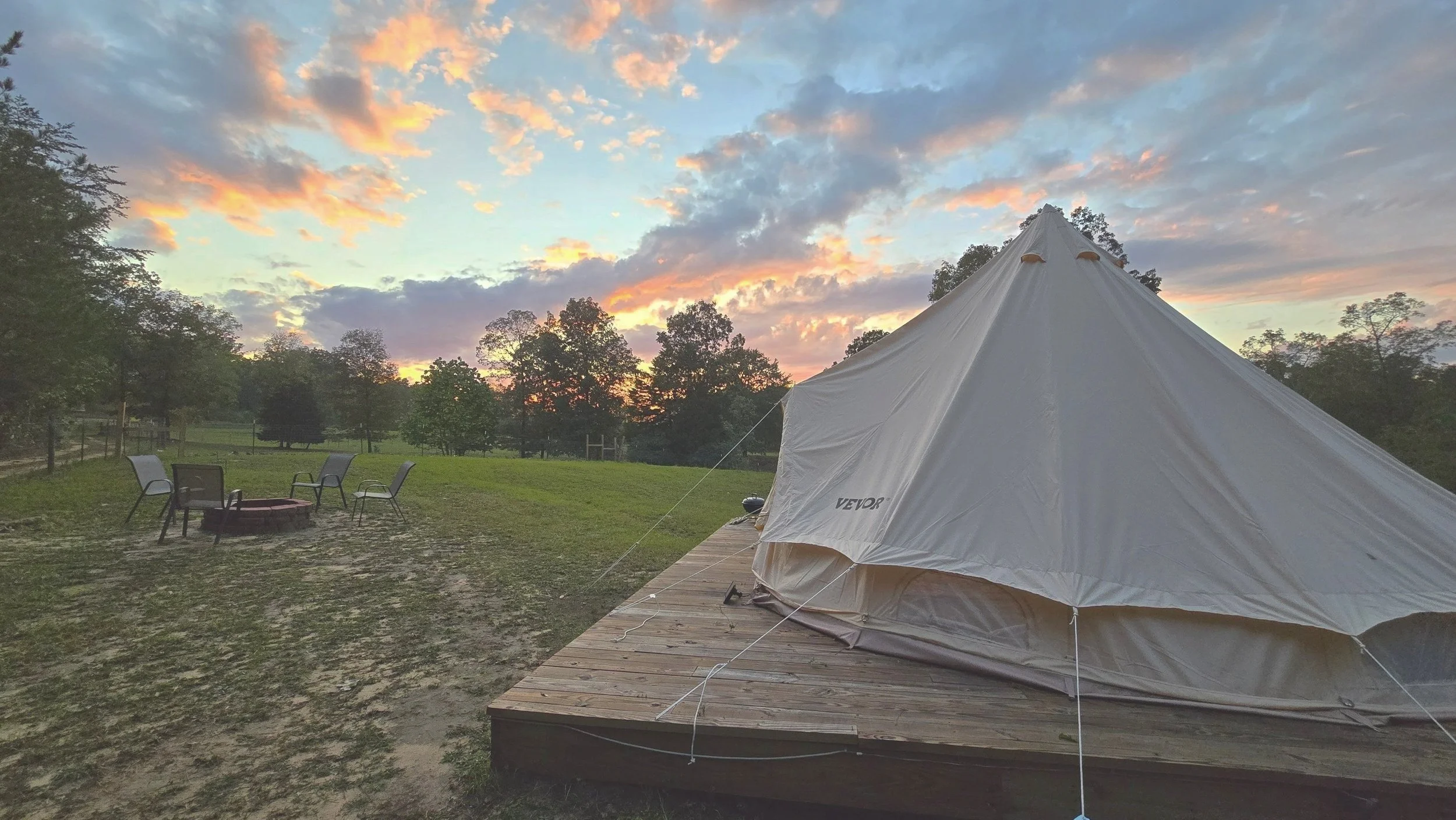 A white teepee tent set up on a wooden platform in an open grassy area during sunset, with a fire pit and four outdoor chairs nearby, and trees and a colorful sky in the background.