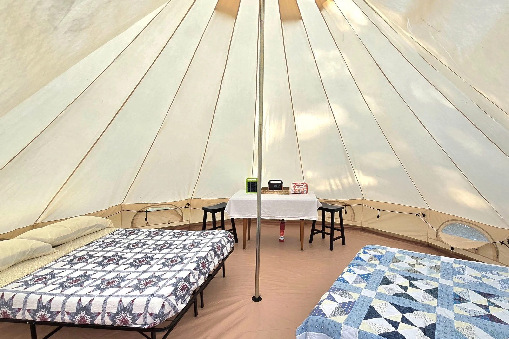 The interior of a large, beige canvas tent with two beds, each with a quilted bedspread, against the walls. A small table with a white tablecloth sits in the center, holding a radio, small solar panel, and a box. Two black stools are placed on either side of the table. There is a fire extinguisher beneath the table, and a string of lights along the tent's perimeter.