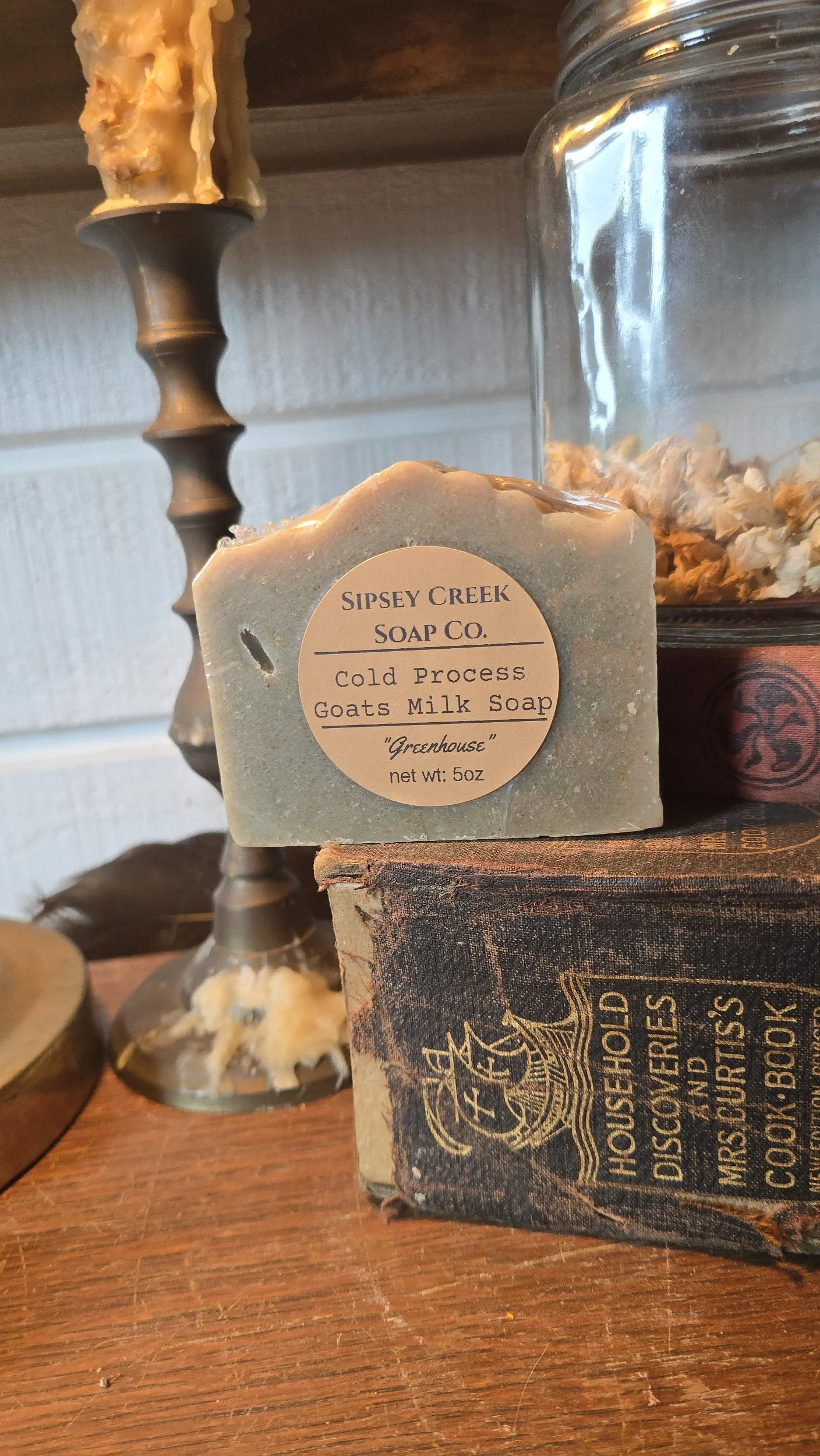 A bar of soap labeled 'Sipsey Creek Soap Co. Cold Process Goats Milk Soap 'Greenhouse'' on a wooden surface with vintage books, a glass jar, and a decorative candle holder in the background.