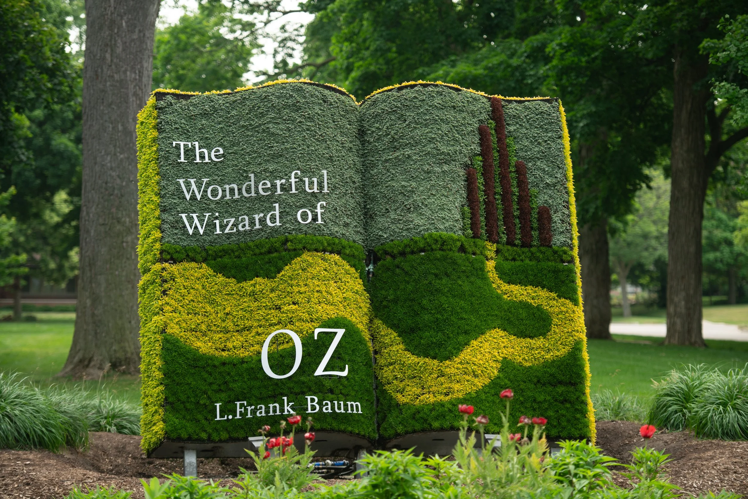 A large, outdoor floral display shaped like an open book, with the title "The Wonderful Wizard of OZ" and the author's name 'L. Frank Baum' on it. The book is decorated with various green, yellow, and brown plants, and is situated in a park with tree