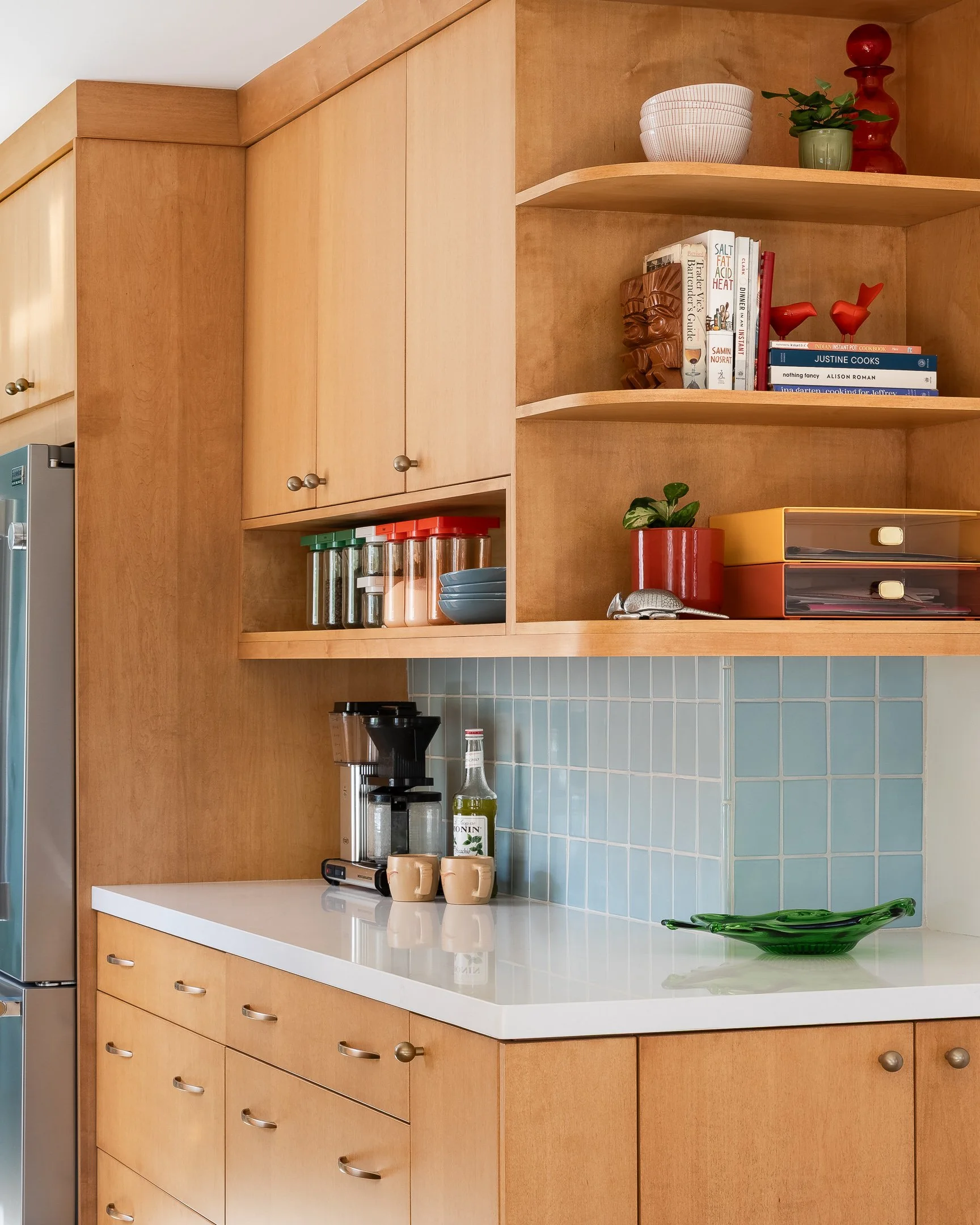 Kitchen with wooden cabinets, a white countertop, blue tile backsplash, coffee maker, bottles, cups, and decorative items such as books, plants, and sculptures.