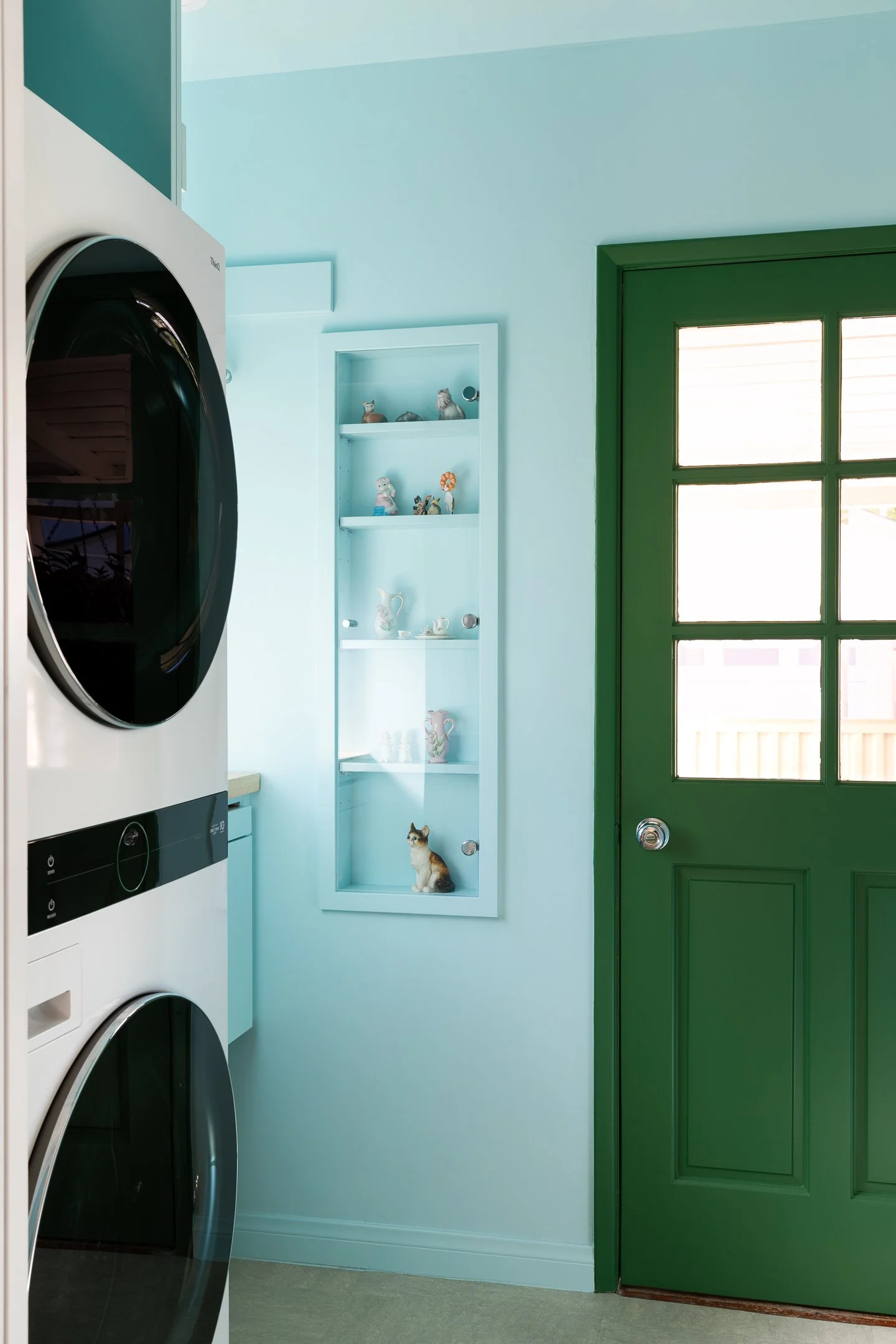 Interior view of a laundry room with a green door and a built-in display shelf with cat figurines and a real cat sitting on the bottom shelf.