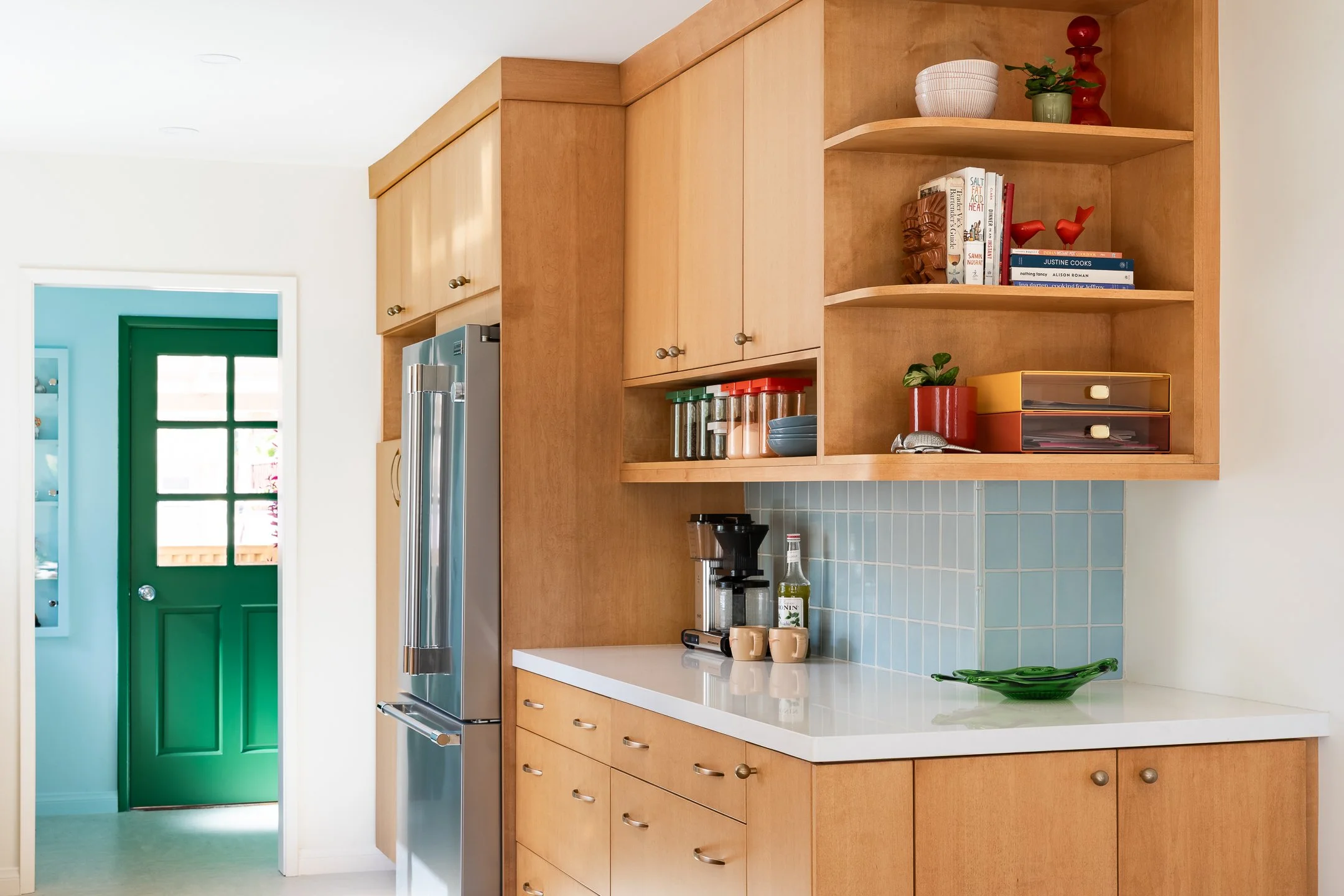 Kitchen with wooden cabinets, a stainless steel refrigerator, blue tile backsplash, and a white countertop with coffee mugs and a coffee maker, adjacent to a green door.